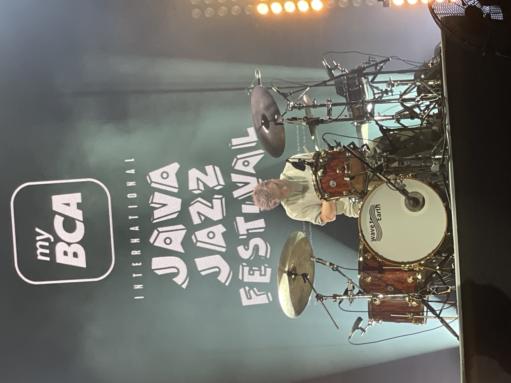 The image depicts a stage during the Java Jazz Festival in Salembaran, Indonesia. A drum set is positioned center-right, with cymbals and a snare drum visible. A drummer with short, light-colored hair is seated behind the drum set. Behind the drummer and the drum set is a large screen displaying the event logo: "my BCA International JAVA JAZZ FESTIVAL." The lighting suggests an indoor venue with stage lights visible at the top of the frame. The scene conveys an active concert environment.