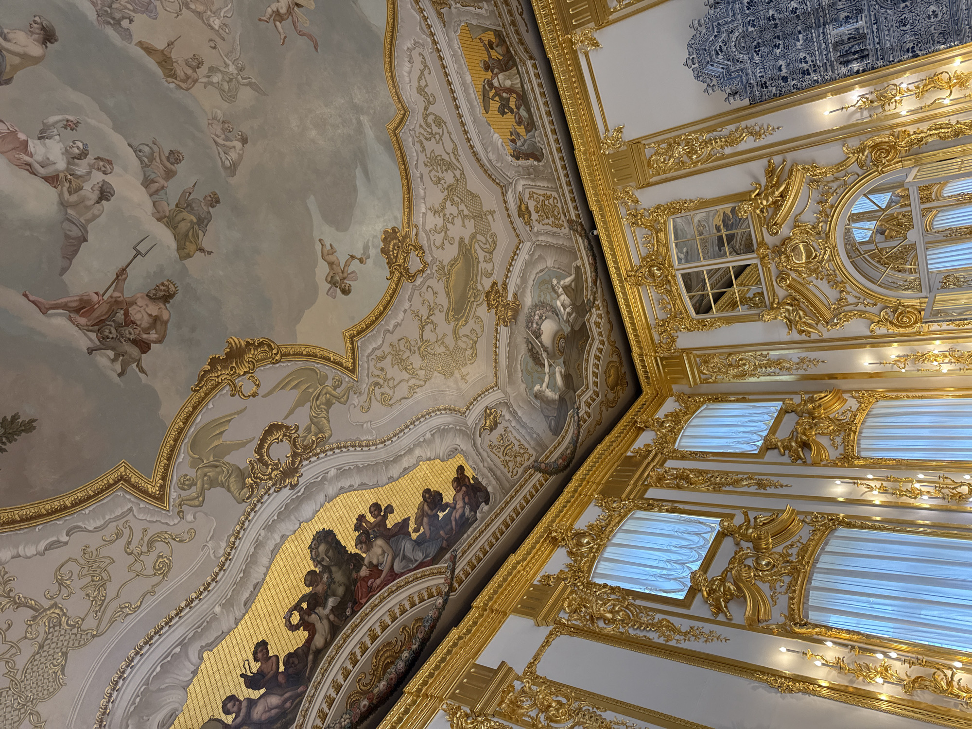 The image depicts the opulent interior of the Catherine Palace in Pushkin, Russia, specifically focusing on the richly decorated ceiling and a portion of the walls. The ceiling is a masterpiece of Baroque art, featuring extensive gilded stucco work and vibrant frescoes. Mythological figures and cherubs are depicted in various poses amidst swirling clouds, with intricate scrollwork and floral motifs adorning the gilded frames. The walls are white and heavily ornamented with elaborate gold carvings, including putti and decorative panels. Several tall windows with sheer white curtains are visible, as well as candelabra mounted on the walls, casting a warm glow. To the right, a section of a blue and white tiled stove or wall panel adds a contrasting element to the predominantly gold and painted decor. The perspective is from below, looking upwards, emphasizing the grandeur and scale of the room.