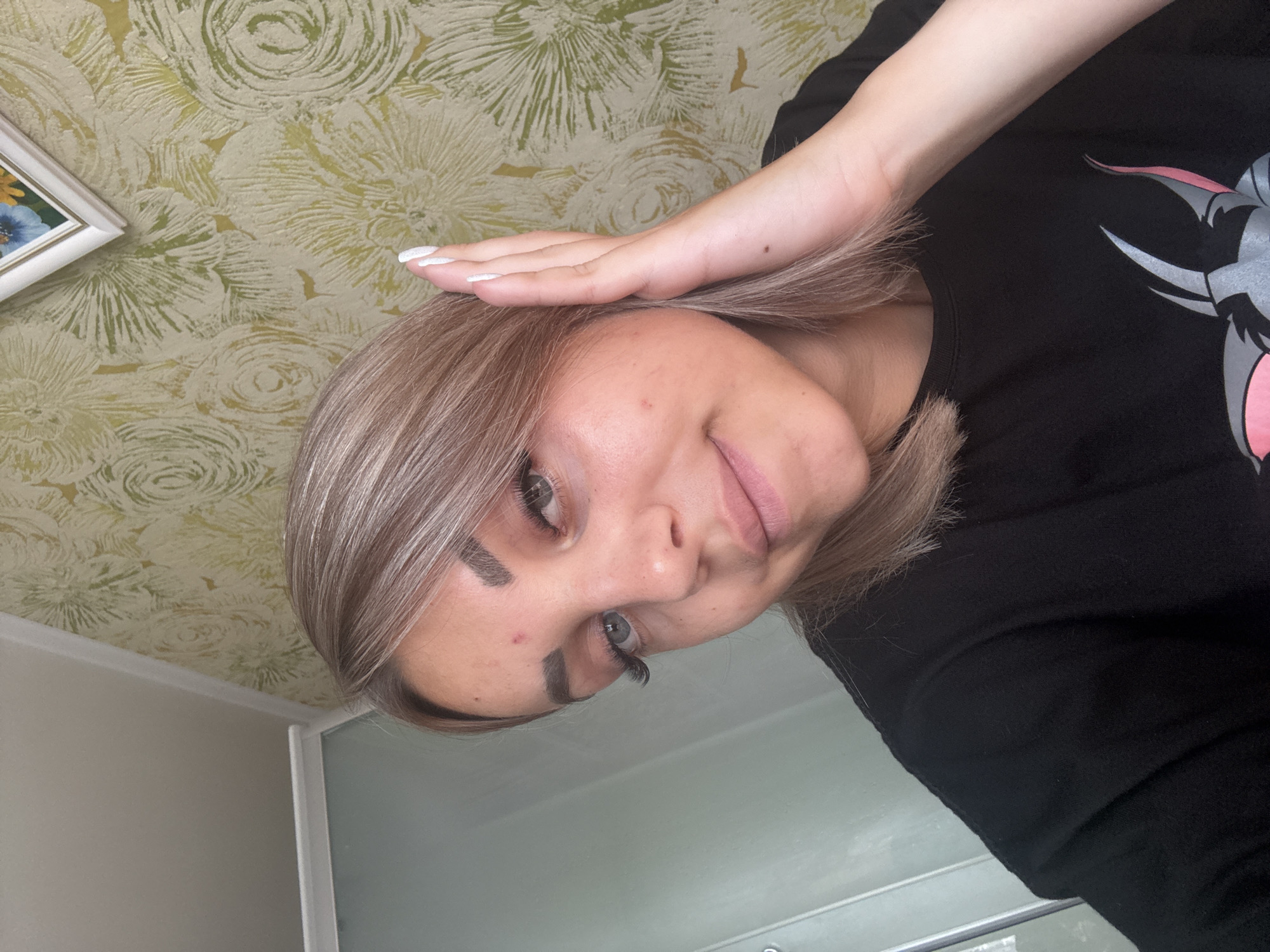 A young woman with blonde hair and blue eyes is captured in a selfie from the chest up. She is wearing a black t-shirt and her left hand is raised to her face, partially obscuring her ear. She has a small pimple on her forehead. The background consists of a wall with a floral pattern and a framed picture. To the left, a portion of a glass shower enclosure is visible. The lighting suggests an indoor setting, likely a bathroom or bedroom. The image was taken in Krasnogorskoye, Russia.