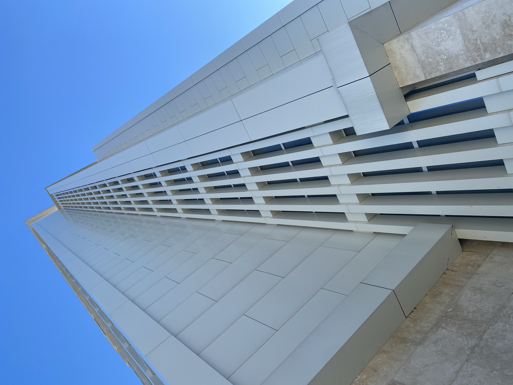 The image is a low-angle shot of a tall, modern building in Sochi, Russia. The building has a predominantly white facade with a pattern of horizontal lines and rectangles. The facade is constructed of rectangular panels with horizontal louvers creating shadow lines, with sections of gray stone at the corners. The design emphasizes verticality. The sky above is clear and blue. There are no people visible in the image.