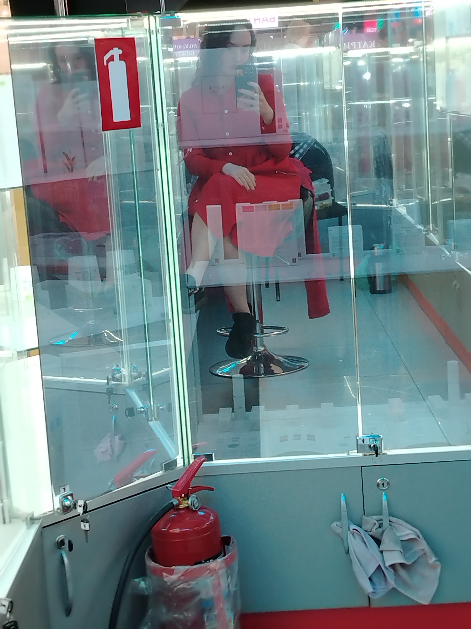 This image shows a retail display case, likely in a shopping center in Izhevsk, Russia, as indicated by the Cyrillic text visible on the glass. Inside the display case, a person is seated on a stool, wearing a red outfit and holding a smartphone. The person appears to be taking a selfie or looking at something on their phone. The display case is made of glass and metal, and it contains various items, including what looks like clothing on hangers and a fire extinguisher on the exterior. Reflections in the glass show more of the shopping center environment, including other display cases and lighting. The overall scene depicts a typical retail environment, with a focus on the individual interacting with their phone within the display.
