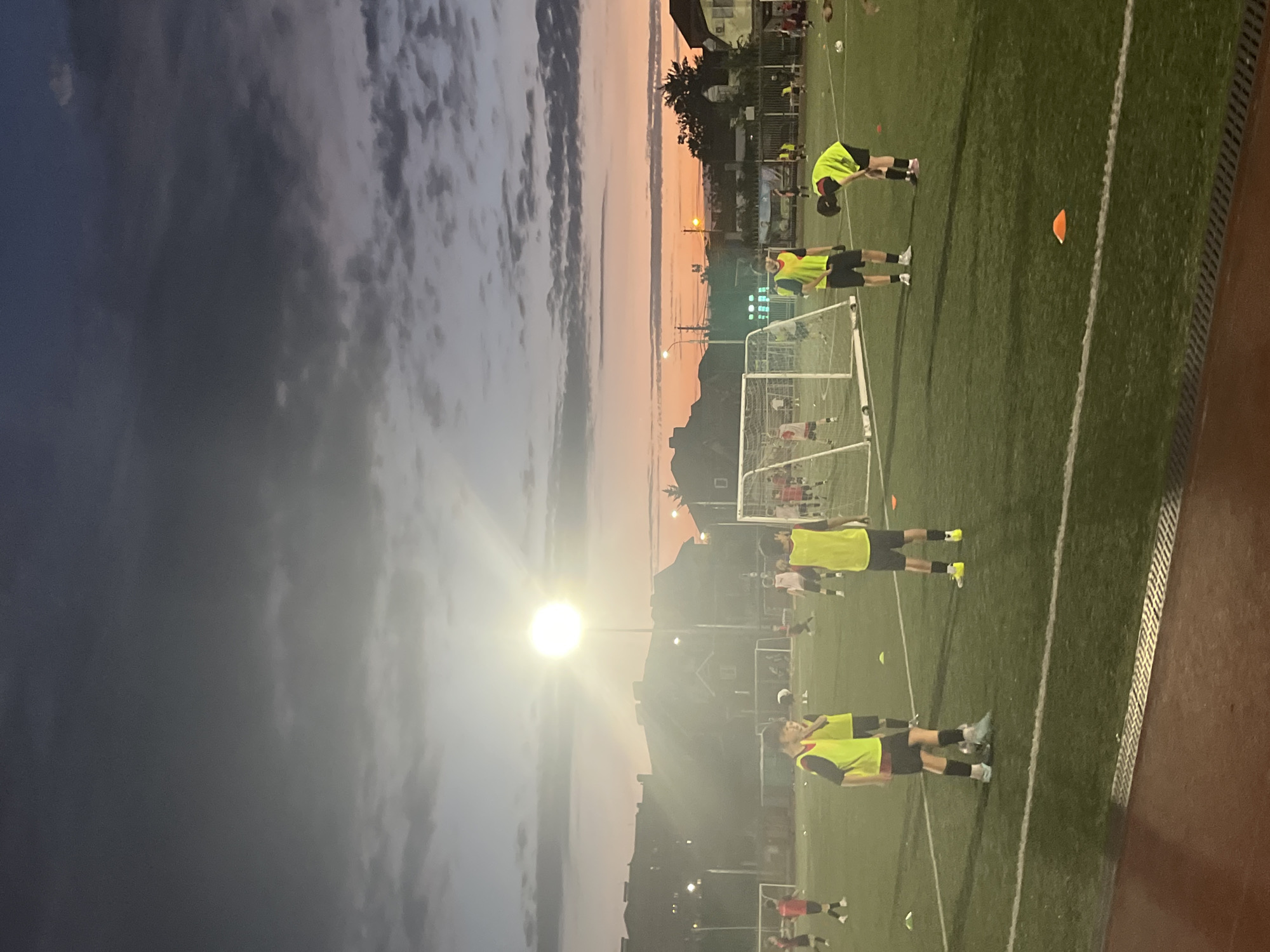 The image shows a soccer practice session on an artificial turf field at dusk in Krasnodar, Russia. Several young males, some wearing bright yellow vests over their athletic attire, are spread across the field. Some are actively engaged in drills, while others stand around. Two soccer goals are visible, one in the foreground and another in the background. The sky above is a gradient of dark gray clouds transitioning into an orange and pink sunset. A bright stadium light illuminates a portion of the field. Residential buildings are visible in the background, suggesting an urban or suburban setting.
