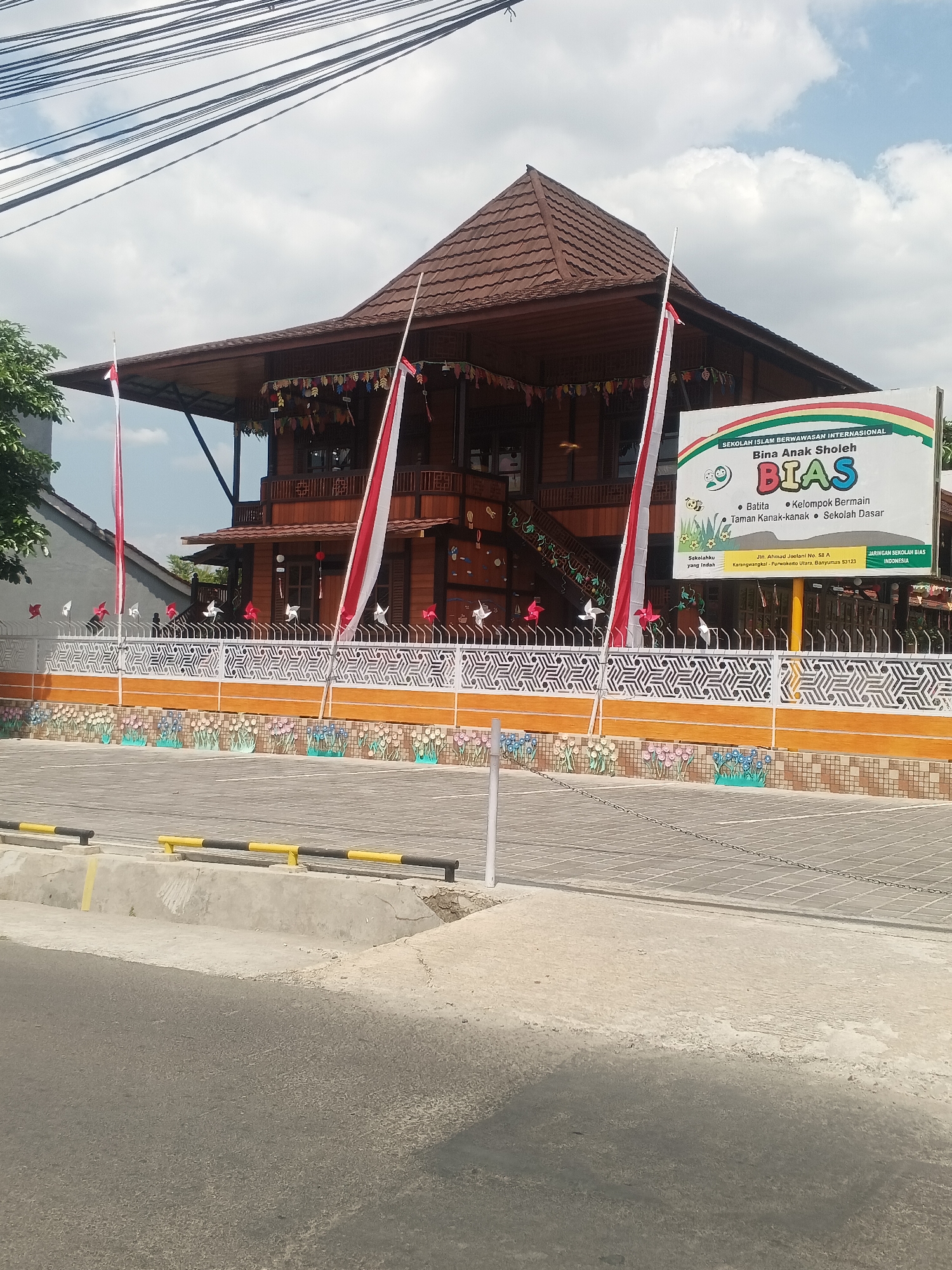 The image shows a three-story wooden building with a brown tiled roof, likely a school or educational institution, situated in Arcawinangun, Indonesia. The building is decorated with small Indonesian flags and colorful pennants along the roofline. In front of the building is a white fence with an orange base and patterned cutouts. The fence is adorned with small red and white pinwheels and miniature flags. A large white sign with text and colorful graphics, including a rainbow and a bee, is prominently displayed to the right of the entrance. The sign indicates "SEKOLAH ISLAM BERWAWASAN INTERNASIONAL" and "Bina Anak Sholeh BIAS," suggesting it is an international Islamic school. The building is surrounded by a paved area with some vegetation visible on the left. A road runs in front of the property, with a concrete barrier and black and yellow striped guardrails visible along the roadside. The sky is partly cloudy.