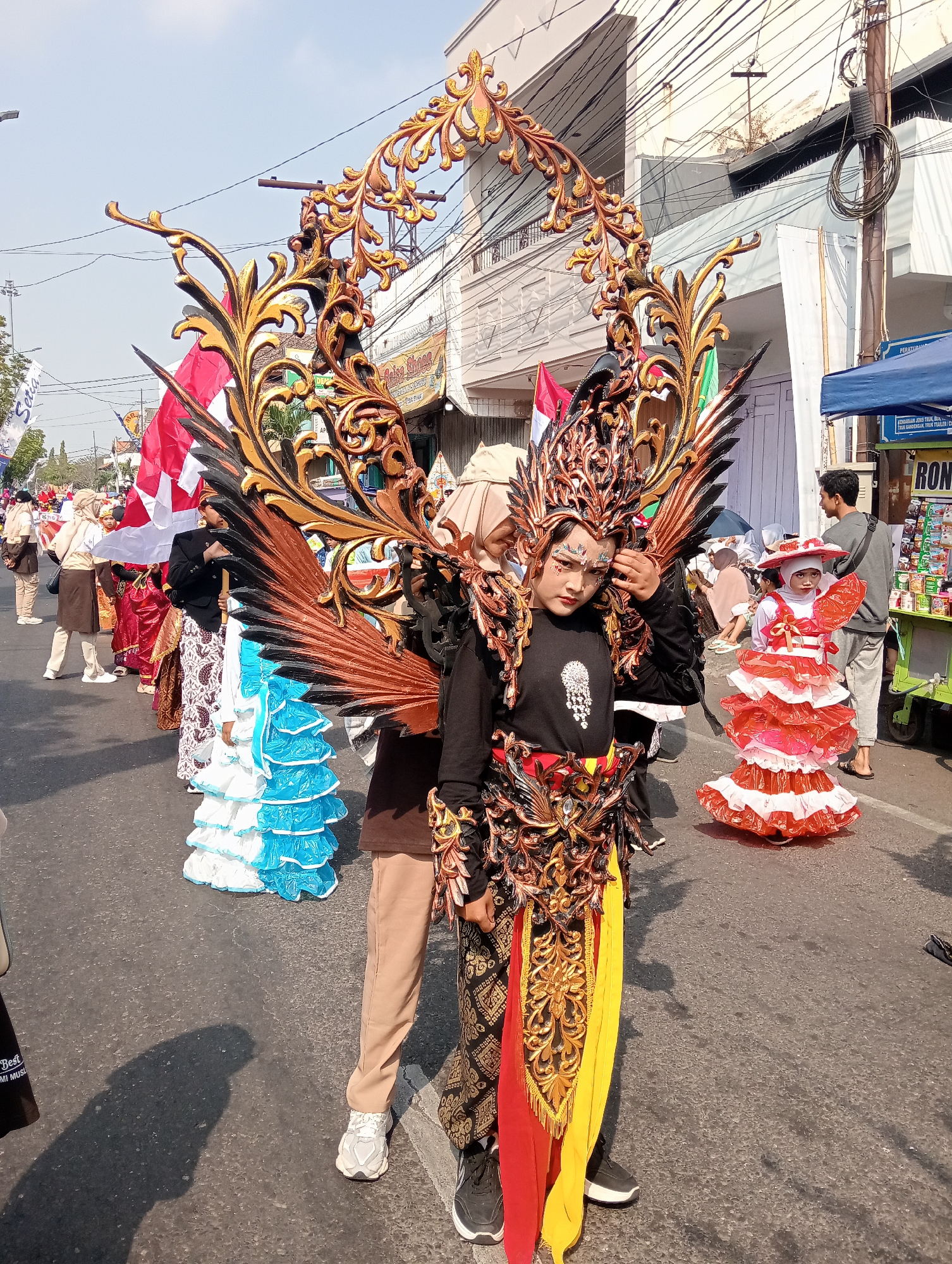 In the city of Bintoro, Indonesia, a street parade is underway. The central figure is a young person in an elaborate costume resembling a mythical creature with large, ornate golden wings and headpiece. The costume features intricate golden detailing on a black base, with draped red and yellow fabric. To the right, a child in a vibrant red and white layered dress and hat stands. In the background, a procession of people dressed in traditional attire, including those in white and blue tiered dresses and a person in a batik skirt, walk along the street. Indonesian flags are visible among the participants. The street is lined with buildings and overhead wires, suggesting an urban setting. A stall selling goods is also present on the right side of the frame. The ground is asphalt, and shadows indicate a sunny day.
