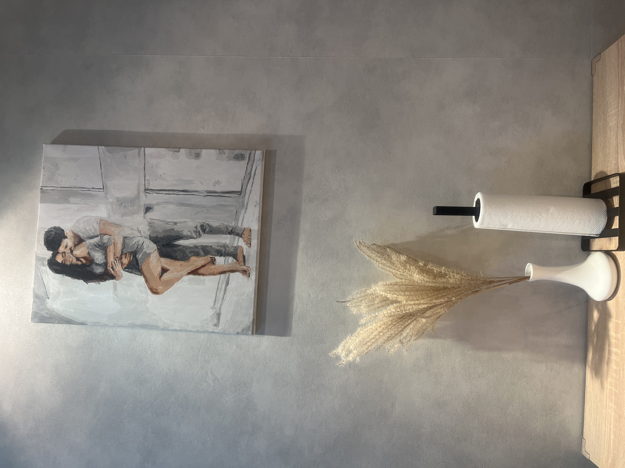 A painting of a couple embracing hangs on a textured gray wall. Below the painting, on a wooden surface, sits a white vase with dried grass and a paper towel holder. The scene is indoors, with soft lighting. There are no people or animals present.