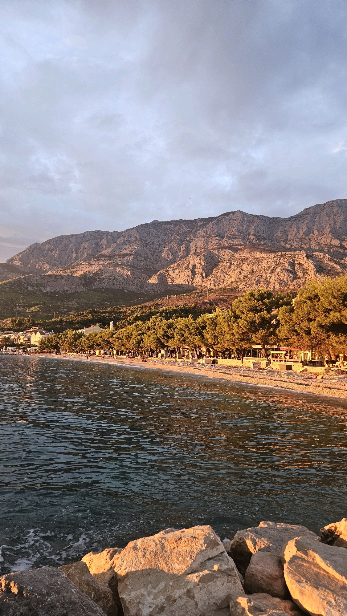 The image depicts a serene beach scene at sunset in Općina Tučepi, Croatia. In the foreground, large, sun-drenched rocks line the edge of the clear, rippling water. Beyond the water, a pebble beach stretches out, dotted with lounge chairs and a few scattered individuals. A line of lush green pine trees provides a natural border between the beach and a row of white buildings with red-tiled roofs. Towering behind the buildings and trees is a majestic mountain range, its rugged slopes illuminated by the warm, golden light of the setting sun. The sky above is a canvas of soft, muted clouds, reflecting the twilight hues.