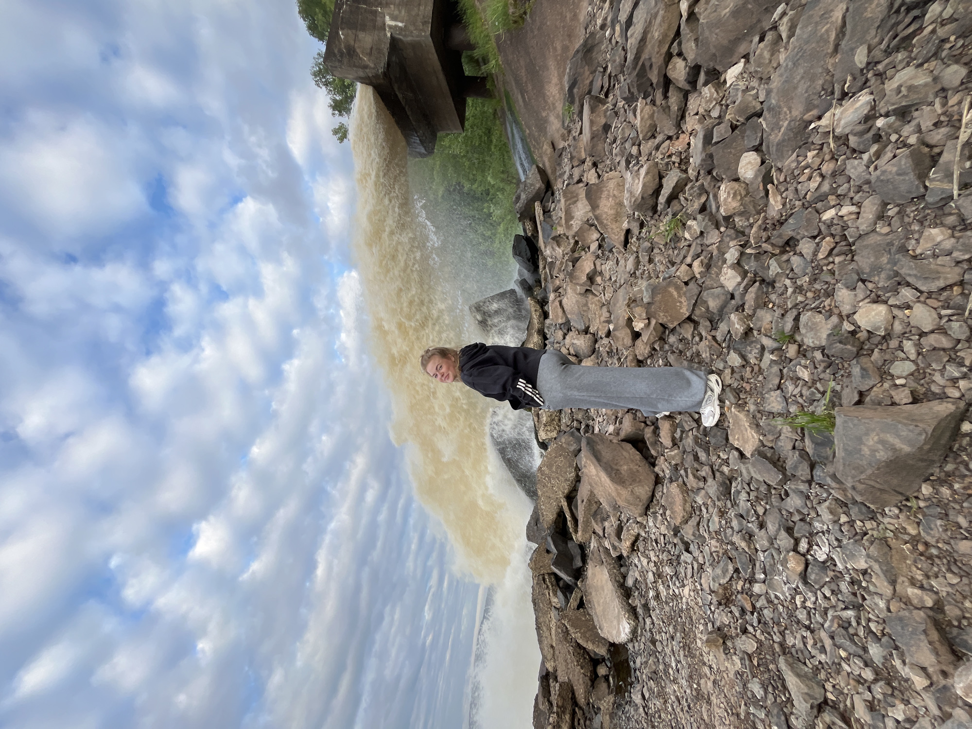 A young blonde woman stands on a rocky shoreline, looking over her left shoulder at the camera. She is wearing a black hooded sweatshirt with white stripes on the sleeves and gray sweatpants. Behind her, a large plume of murky water is being forcefully discharged from a concrete structure. The sky is filled with dramatic, cloudy formations. The surrounding area is covered in various sizes of rocks and pebbles, with some sparse green vegetation visible on the right side of the frame. The location is identified as Chernushka, Russia, though specific landmarks are not discernible in the image to pinpoint an exact spot within the city. The overall scene suggests a potentially industrial or infrastructural water release in an outdoor, somewhat rugged environment.