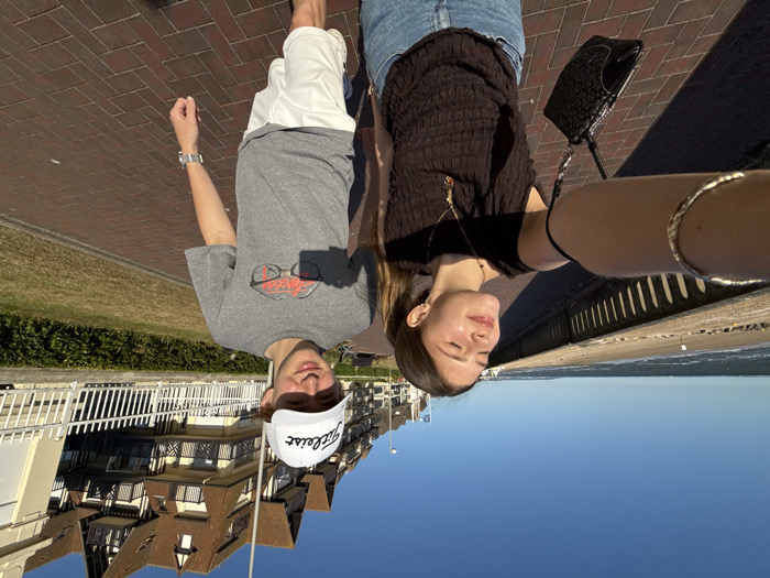 A selfie shows two people walking outdoors. A man wearing a gray t-shirt and white hat with a logo walks alongside a woman in a black shirt and jeans. The background includes a brick walkway, a building with a Tudor-style facade, a green strip of grass, a beach, and the sky. The image appears to be taken sideways, with the sky at the bottom and the building at the top.