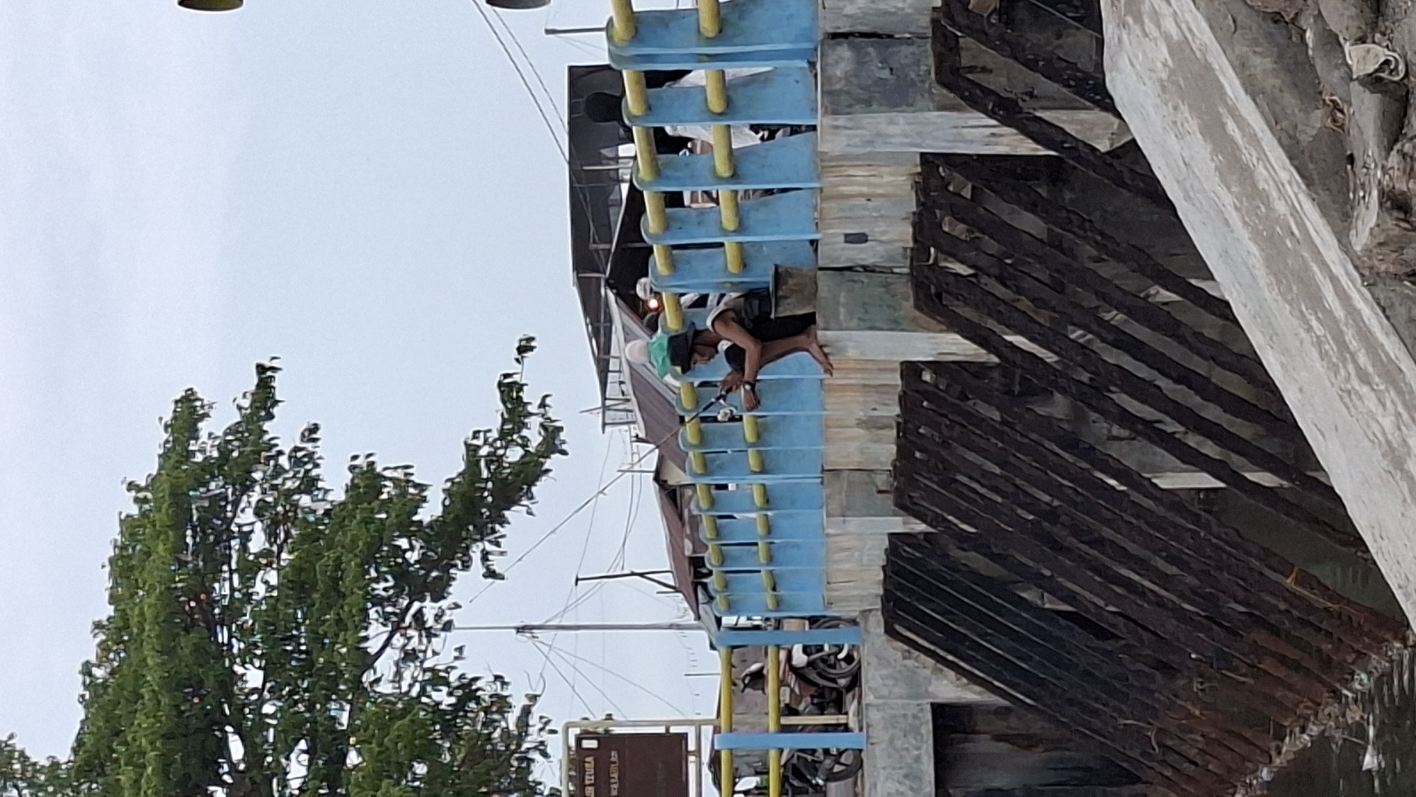 A person is fishing on a bridge in Lhokseumawe, Indonesia. The person is wearing a black hat, a light-colored tank top, and dark shorts, and is sitting on the edge of the bridge with their legs dangling. They are holding a fishing rod and appear to be focused on the water below. The bridge has blue railings with yellow accents and is made of concrete. In the background, there are buildings with corrugated metal roofs, and a tree with green leaves overhangs the scene. There is also a motorcycle parked on the left side of the bridge. The overall scene suggests a casual, everyday activity taking place in an urban or semi-urban environment near a body of water.