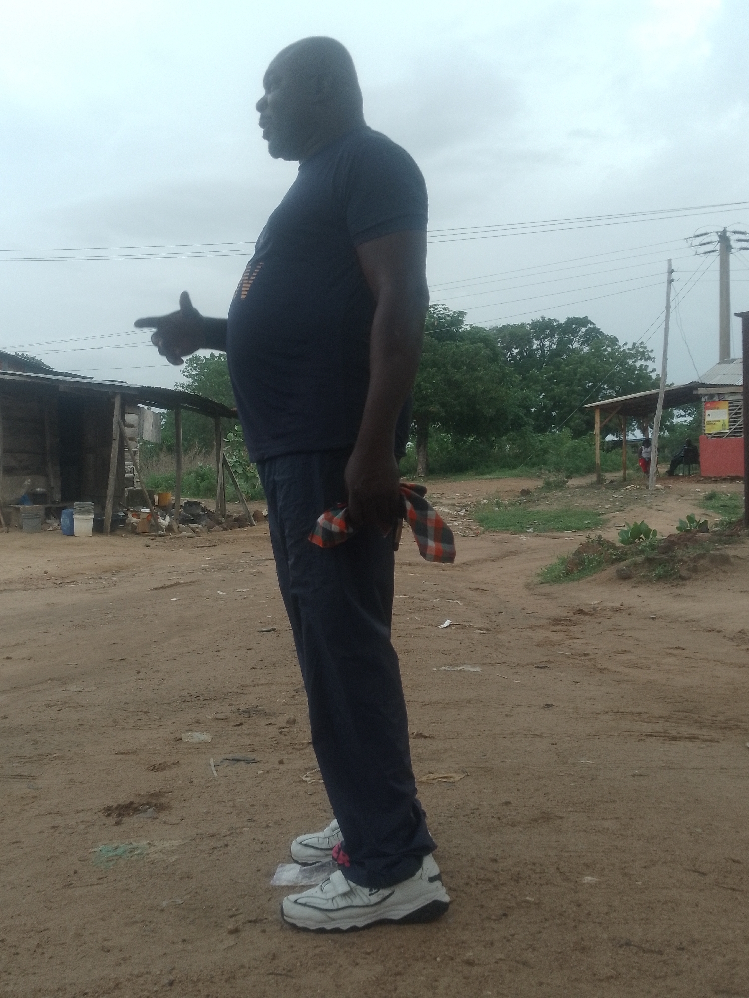 An eye-level, full shot depicts a man in Adankolo, Nigeria, standing in a sandy, dirt area, in front of various buildings and trees under an overcast sky. The man, dark-skinned with a bald head, faces left, pointing his left hand outward. He wears a dark blue short-sleeved shirt and dark blue sweatpants, coupled with white sneakers. He holds a small red and gray checkered cloth in his right hand.
In the background, to the left, there is a rickety, wooden building with a tin roof; chickens roam near it. Further back and to the right is a patch of green trees, while a small, red-painted structure with a canopy stands in the distance. A utility pole is also visible. In front of the man, the ground is mostly dirt and sand, with some scattered debris.