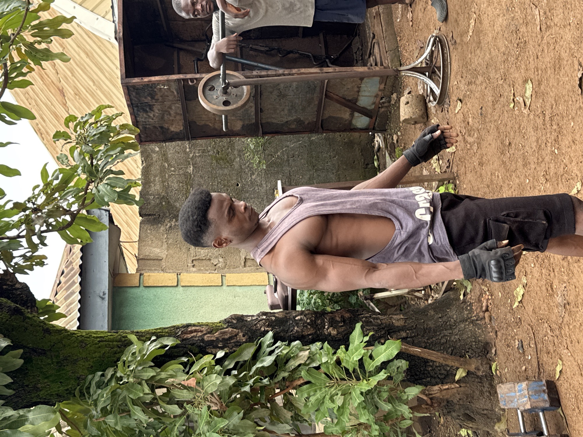 The image captures a young man with a muscular physique in a dimly lit outdoor gym setting, possibly in Akwanga, Nigeria, given the context provided. He is wearing a grey tank top, black shorts, and black fingerless gloves, and appears to be engaged in a workout. His arms are flexed, showcasing his developed muscles. To his right, another man stands near a weightlifting rack, leaning on the bar. The environment is rustic, with a dirt ground, a large tree with moss on its trunk on the left, and corrugated metal roofing visible in the background, suggesting a developing or informal urban setting. The overall atmosphere is one of determination and physical exertion amidst a humble workout space.