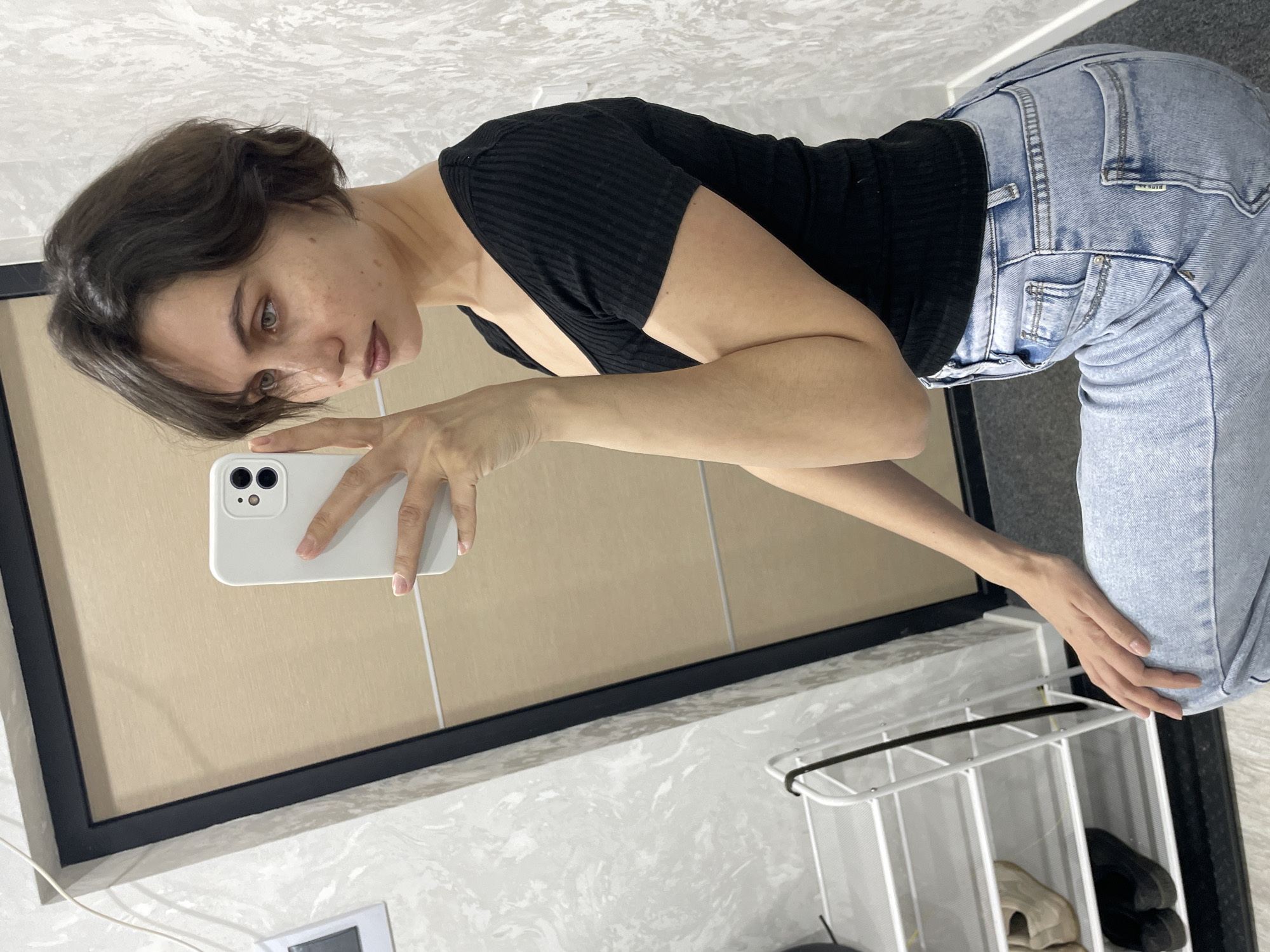 A woman is taking a selfie in a bathroom. She is wearing a black ribbed top and light blue jeans. She is holding a white smartphone in her left hand. The bathroom has light-colored tile and a black-framed mirror. A white shoe rack with shoes is visible. No specific location within Izhevsk, Russia can be determined from the image.