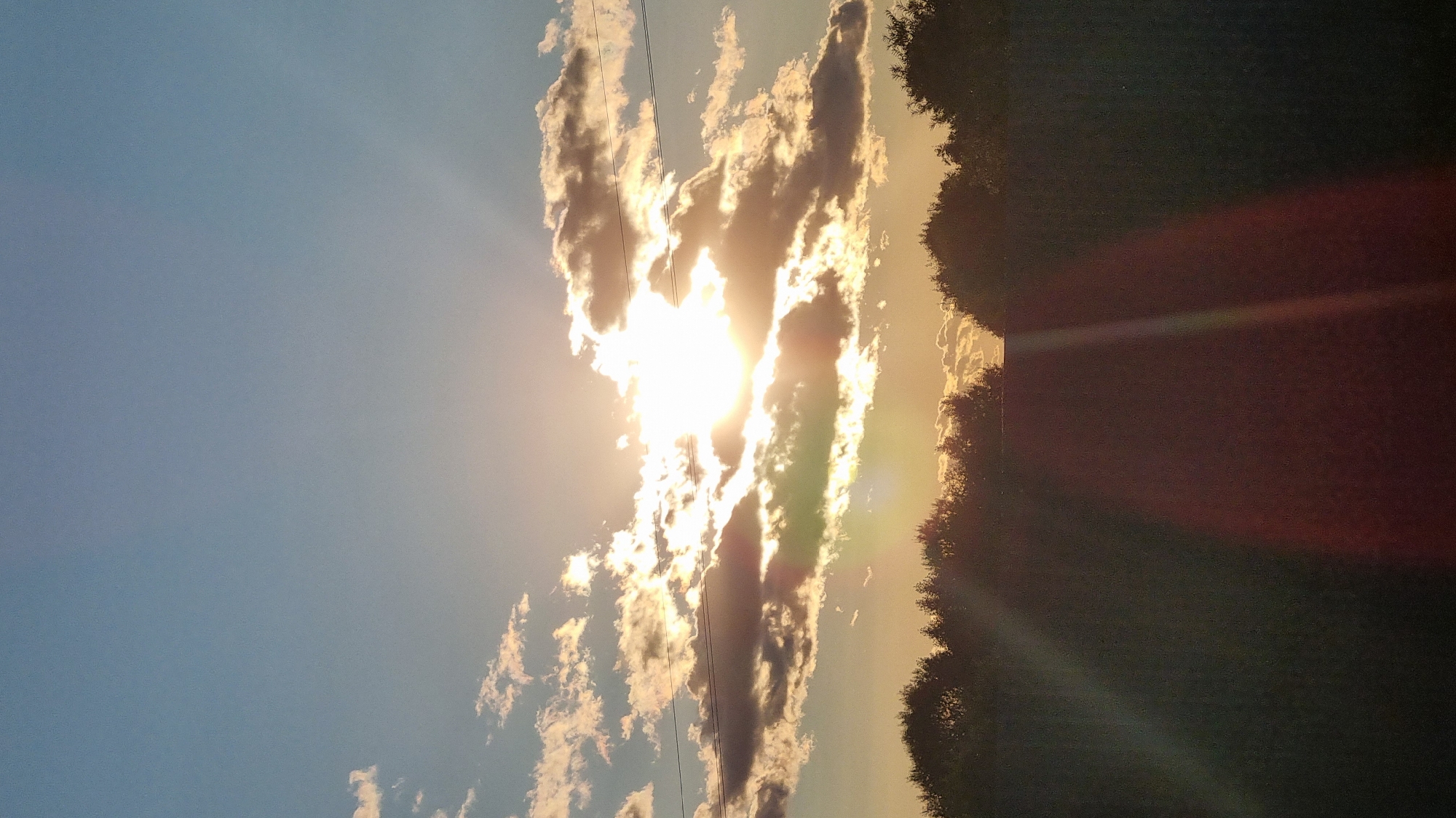 A sunset with the sun peeking through clouds is captured over a line of trees. The sky is a clear blue with scattered white and gray clouds, some of which are illuminated by the sun's rays. A faint power line is visible in the sky, stretching horizontally. Below the trees, a dark, indistinct foreground suggests a field or landscape. The image appears to be a photograph taken outdoors in the city of Vysokoe, Russia.