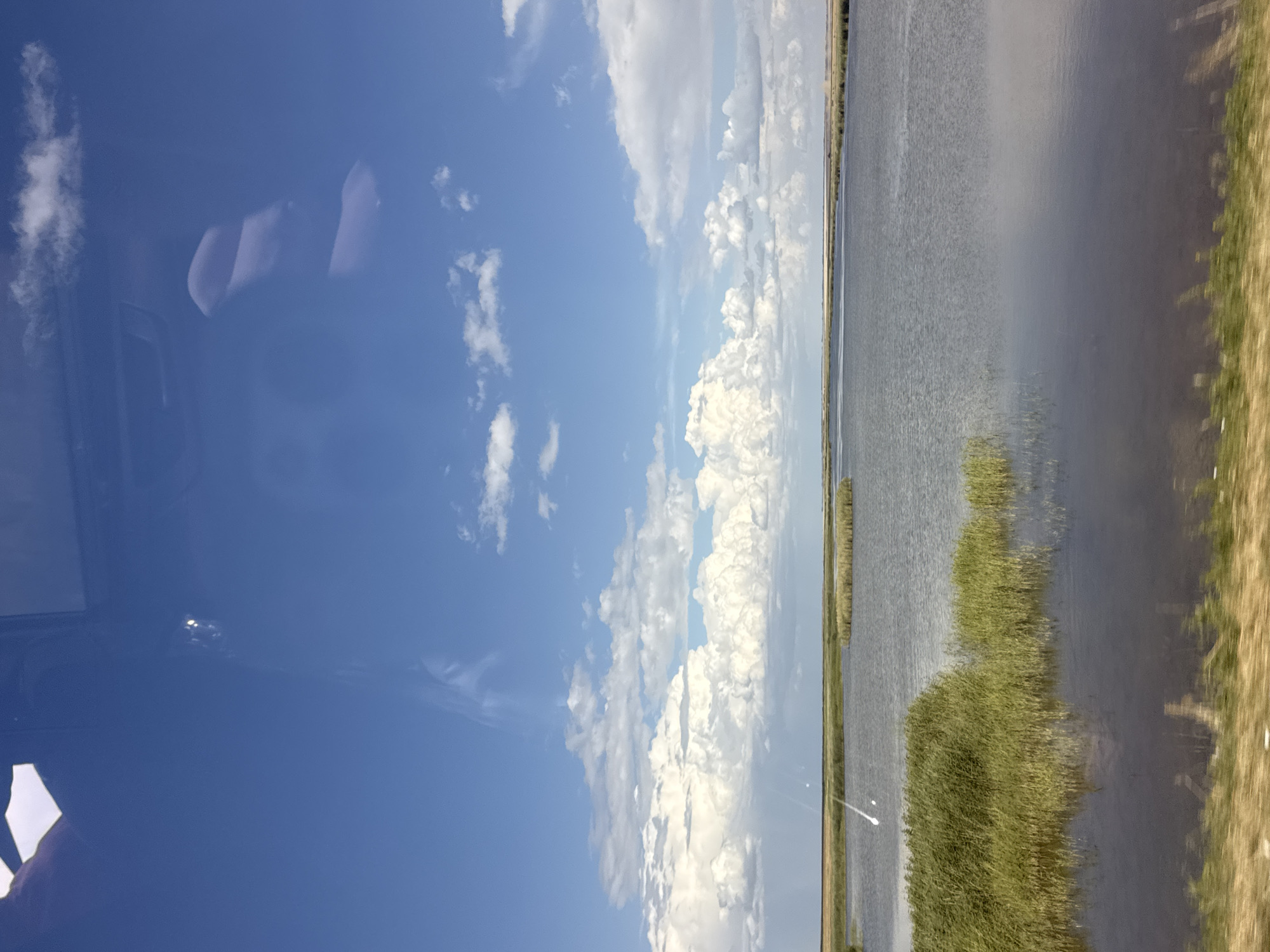 The image displays a vast, calm body of water, likely a lake or slow-moving river, under a bright blue sky filled with puffy white clouds. The water's surface is mostly still, reflecting the sky and creating a slightly hazy, serene atmosphere. Along the right edge of the water, tall, green reeds or marsh grass grow densely, partially obscuring the land beyond. To the far right, a strip of dry, yellowish grass suggests a shoreline or embankment.
In the upper left quadrant of the image, reflections on a smooth surface, possibly a window, reveal blurry shapes. These shapes appear to be interior elements of a vehicle, with what could be the headrests of seats and a portion of a dashboard or window frame visible. The reflection also shows distorted images of clouds and the blue sky.
The overall scene suggests a peaceful, outdoor setting, possibly experienced from within a vehicle or dwelling with a large window overlooking the water. The bright sky and abundant clouds indicate daytime, likely mid-morning or afternoon, with fair weather. The location cues, such as the marshy vegetation and the calm water, are consistent with a rural or natural landscape. No text is visible in the image.