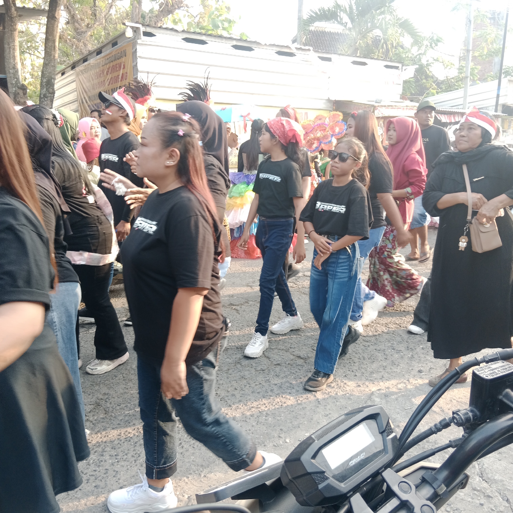 In the city of Tegalrejo, Indonesia, a group of people are participating in a parade. The foreground is dominated by the handlebars and digital display of a motorcycle, suggesting it is part of the procession or being used by a photographer.
A diverse group of individuals are visible, many wearing black t-shirts with a white logo that reads "VAPER." Some are also wearing red and white bandanas, and one person sports a red and white baseball cap. The participants appear to be of various ages and genders.
The backdrop consists of a street with a building on the left, possibly a shop or roadside stall, featuring a banner with text in Indonesian. Trees with foliage are present on the left and right, indicating an outdoor setting. The overall atmosphere suggests a festive event, possibly a cultural celebration or a local parade. The lighting indicates it is daytime, with the sun casting shadows.