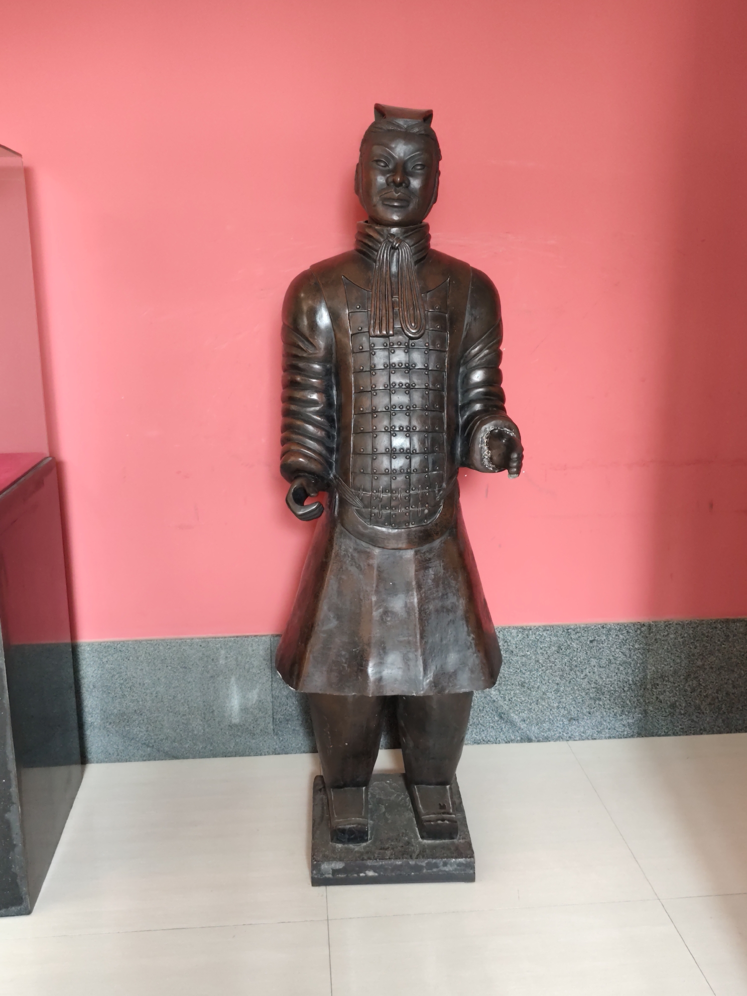 The image shows a full-body bronze statue of a Terracotta Warrior standing against a plain pink wall. The statue is approximately life-sized and depicts a figure in armor with a skirt. The armor has intricate detailing, with rows of small circles or studs. The figure wears a headdress and has a stern facial expression. The statue is mounted on a dark rectangular base, which is placed on a light-colored tiled floor. To the left of the statue, a portion of a dark object, possibly a display case, is visible. The wall behind the statue has a granite-patterned strip along the bottom. The overall environment suggests an indoor setting, likely a museum or exhibition space, potentially in 幸福乡, China, given the stylistic resemblance to the Terracotta Army.