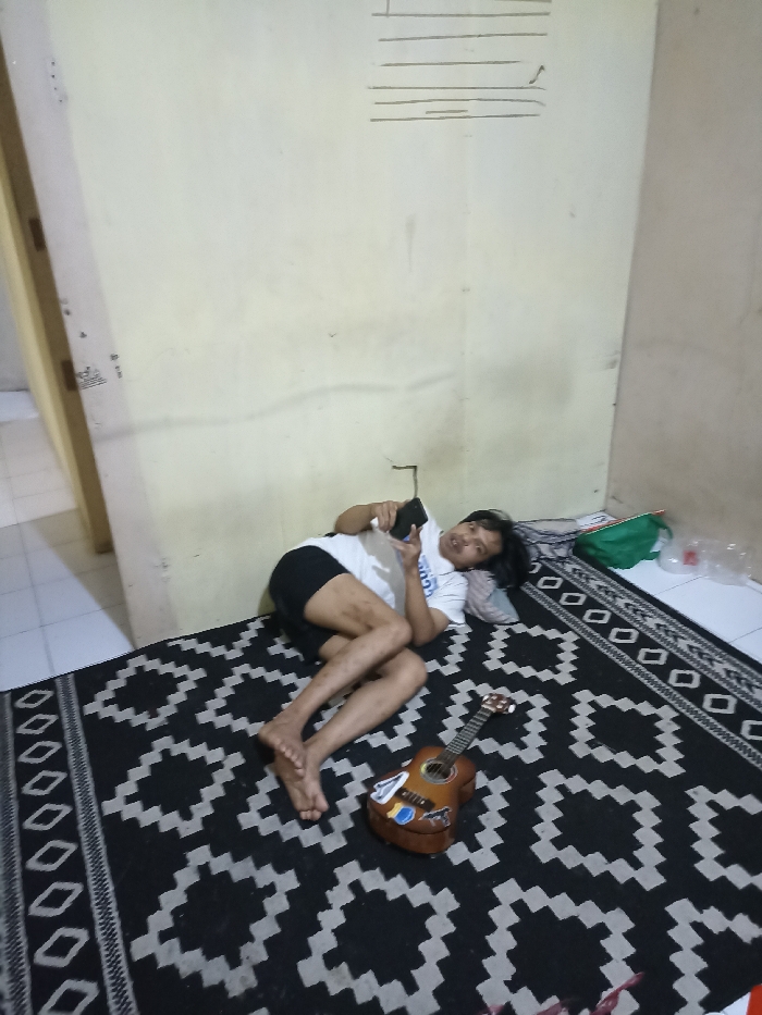 The image shows a man lying on his back on a black and white patterned rug in a room in Jatinangor, Indonesia. He is holding a smartphone in his right hand, looking at it, and talking. His legs are extended, and his right foot is touching the rug. A brown ukulele lies on the rug beside him. There is a green bag in the upper right of the image and white plastic containers on the floor nearby. The room has white walls and is partially visible. The man appears to be in his late teens or early twenties and is wearing a white T-shirt and dark shorts. The room is brightly lit.