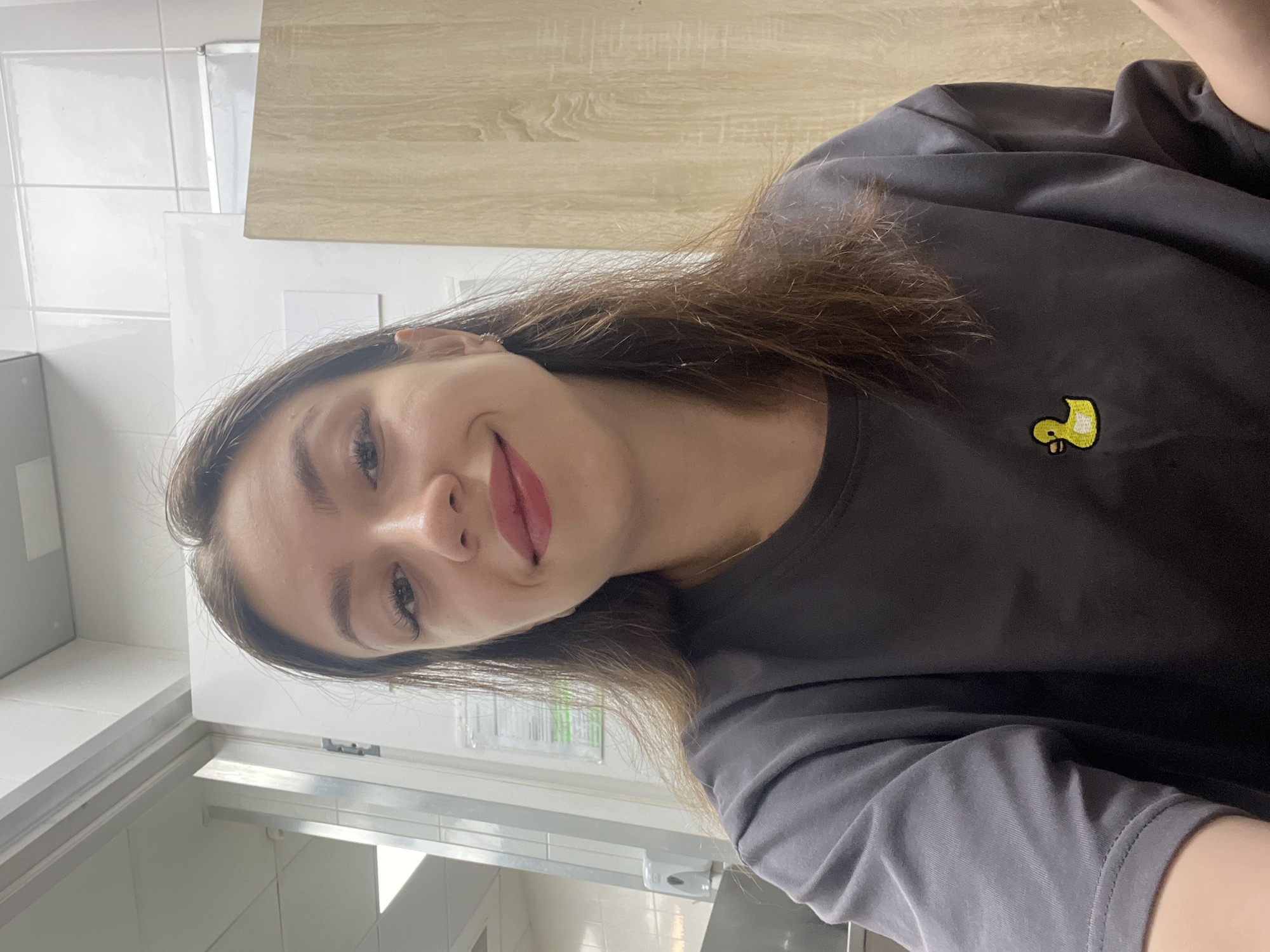 A young woman with long brown hair and fair skin is taking a selfie in what appears to be a public restroom or a similar utilitarian indoor space. She is wearing a dark gray t-shirt with a small embroidered yellow duck on the left side of the chest. Her makeup includes mascara and lipstick. In the background, there is a tiled wall, a wooden door or cabinet, and some shelving or fixtures. The overall lighting suggests an indoor, possibly commercial or institutional setting. The city of Chaikovsky, Russia, is not visually represented in this image.
