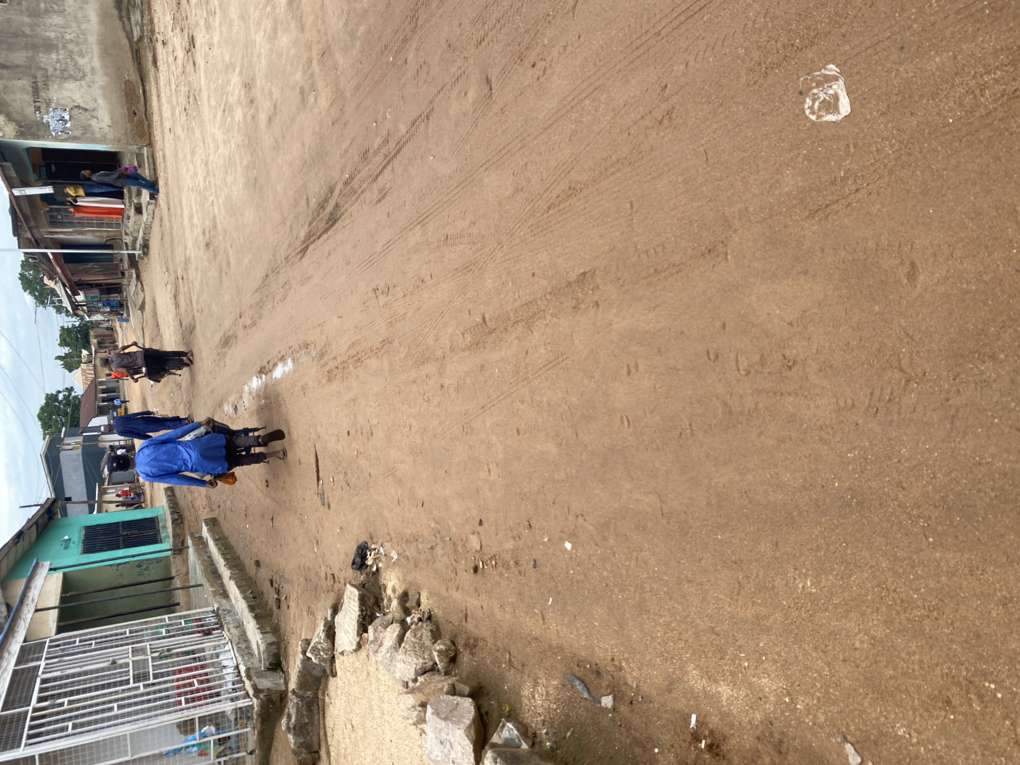 This image depicts a street scene in Jalingo, Nigeria, with a focus on the unpaved, dusty road surface marked by tire tracks. The scene is likely taking place during the daytime, given the visible shadows and the overall brightness of the environment. The weather appears to be dry and possibly warm, as indicated by the dust and the open nature of the setting.
Several people are present in the image. In the mid-ground, a person wearing a bright blue garment is walking away from the camera, pulling a cart. Another person in dark clothing is walking beside them. Further in the background, near some buildings, a few individuals can be seen. One person is standing near the entrance of a building, while others are partially visible.
The setting includes buildings lining the side of the street, some of which appear to be shops or homes. One building is painted a distinctive teal color and has barred windows. Another structure on the left has a more weathered concrete facade with some posters or notices attached. In the foreground, there is rubble and debris, including rocks and some scattered trash. A white plastic bag is also visible on the road surface further back.
The overall impression of the scene is one of everyday life in a developing urban or semi-urban area, characterized by unpaved roads, modest buildings, and pedestrian activity. There is no clear indication of a specific event, but rather a snapshot of a typical day. The lack of distinct landmarks makes precise location identification difficult beyond the provided context of Jalingo, Nigeria. There is some illegible text on the building to the left.
