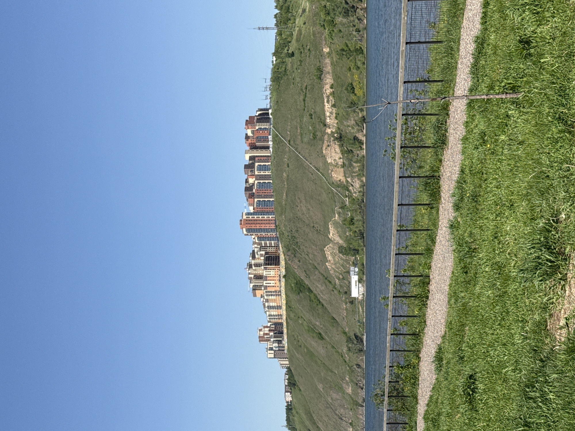 This media file presents a panoramic landscape view of a city riverside, likely Krasnoyarsk, Russia, under clear blue skies during bright daylight. The image is oriented vertically but should be viewed horizontally.
In the foreground, a vibrant expanse of green grass dominates the right side of the frame, bordering a light-colored gravel path that meanders horizontally. To the left of the path, a dark metal fence runs parallel, separating the path from a tranquil body of blue water, characteristic of a river. Scattered small branches and green foliage are visible near the fence, some extending over the water.
Across the water, a steep, green hillside rises, covered with lush vegetation. Along the upper ridge of this hill, a long stretch of multi-story residential buildings can be seen. These modern structures exhibit a variety of warm-toned facades, including terracotta, beige, and white, with numerous windows. They form a prominent skyline against the natural backdrop. Further into the distance on the left, the hillside continues, with more indistinct buildings and possibly a power line tower visible on a higher elevation. A small, cubic white building is also discernible closer to the water on the distant bank.
The sky is uniformly clear and light blue, indicating fair weather. There are no people visible, and no discernible activity is taking place, presenting a serene urban-natural vista. No text is visible in the image.