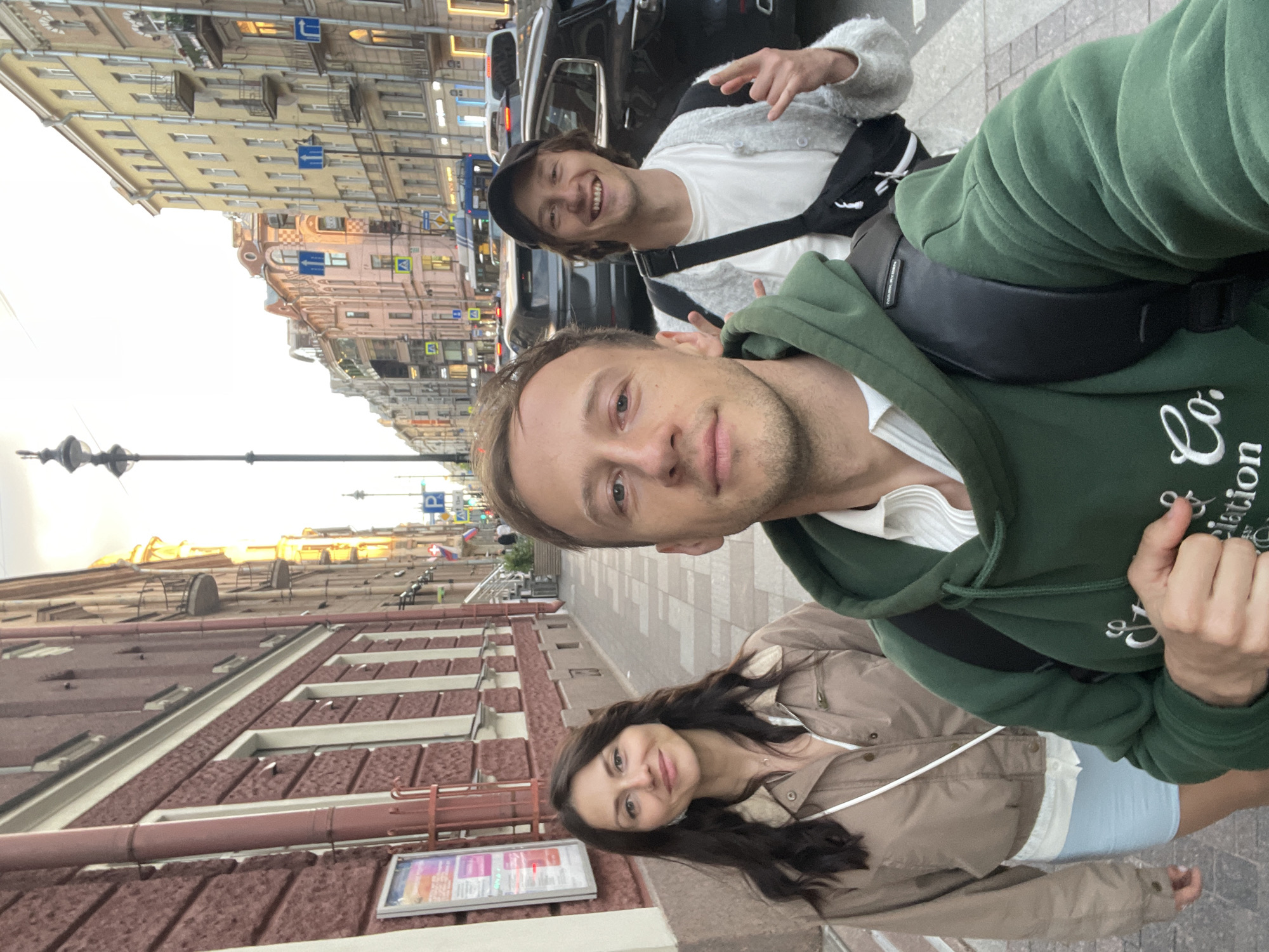 Three individuals are captured in a selfie-style photograph on a city sidewalk, appearing to be in Saint Petersburg, Russia, given the architectural style of the buildings. In the foreground, a man in a dark green hoodie with a white collared shirt underneath is holding the camera. To his left is a woman with long, dark hair, wearing a light brown jacket over a white top and a light blue skirt. To the right of the man in the foreground, another man is visible, wearing a black baseball cap, a white t-shirt, and a light gray cardigan. He is also wearing a black crossbody bag and has his hands raised in a gesture. Behind the individuals, a street scene unfolds with cars and a blue bus on the road, and multi-story buildings with balconies lining the sidewalk. The buildings exhibit characteristic Saint Petersburg architecture, with ornate facades and distinct window designs, suggestive of a historic urban district. The sky appears overcast, indicating likely twilight or a cloudy day.