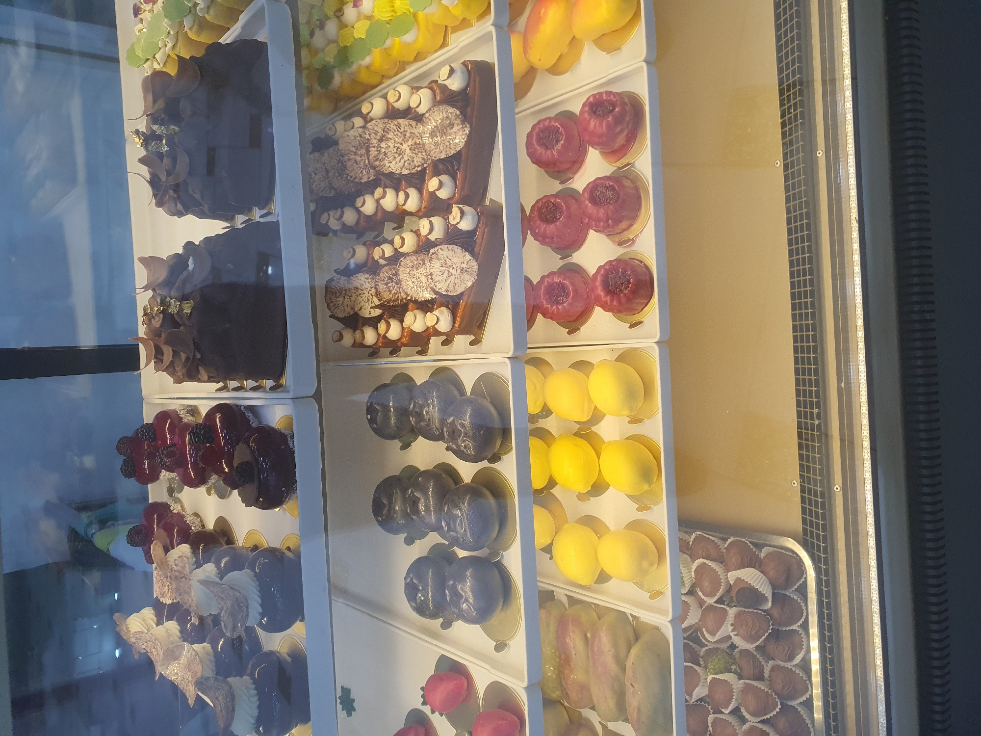 The image displays a refrigerated display case filled with an assortment of pastries and chocolates, likely in a bakery or patisserie in Fergana City, Uzbekistan. The display case is divided into multiple shelves, showcasing a variety of meticulously crafted desserts.
On the top shelf, there are several dark chocolate cakes topped with decorative elements, including what appear to be chocolate shavings and gold leaf. Next to these, to the right, are rows of small, bright yellow pastries, some adorned with green accents. To the left of the chocolate cakes are smaller, dark-colored individual desserts topped with berries and a swirl of cream, some with an accompanying decorative element resembling a fan.
The middle shelf features rows of round, dark purple desserts, with three in the front row. To their left, on the far left of this shelf, are two heart-shaped pink pastries. To the right of the purple desserts, there is a tray of elongated, pale-colored pastries, some with a pinkish hue, and further to the right, several yellow, lemon-shaped pastries are arranged on gold bases.
The bottom shelf contains a tray filled with numerous small, individually wrapped chocolates and what appear to be chocolate-covered strawberries. Some of the chocolates are nestled in white paper liners. To the right of the chocolates, there are two mango-shaped pastries on gold bases, and to the right of those, there are additional yellow, lemon-shaped pastries.
The lighting in the display case highlights the glossiness of the desserts. The overall environment suggests a commercial setting focused on high-end confectionery. There are no visible people in the image.