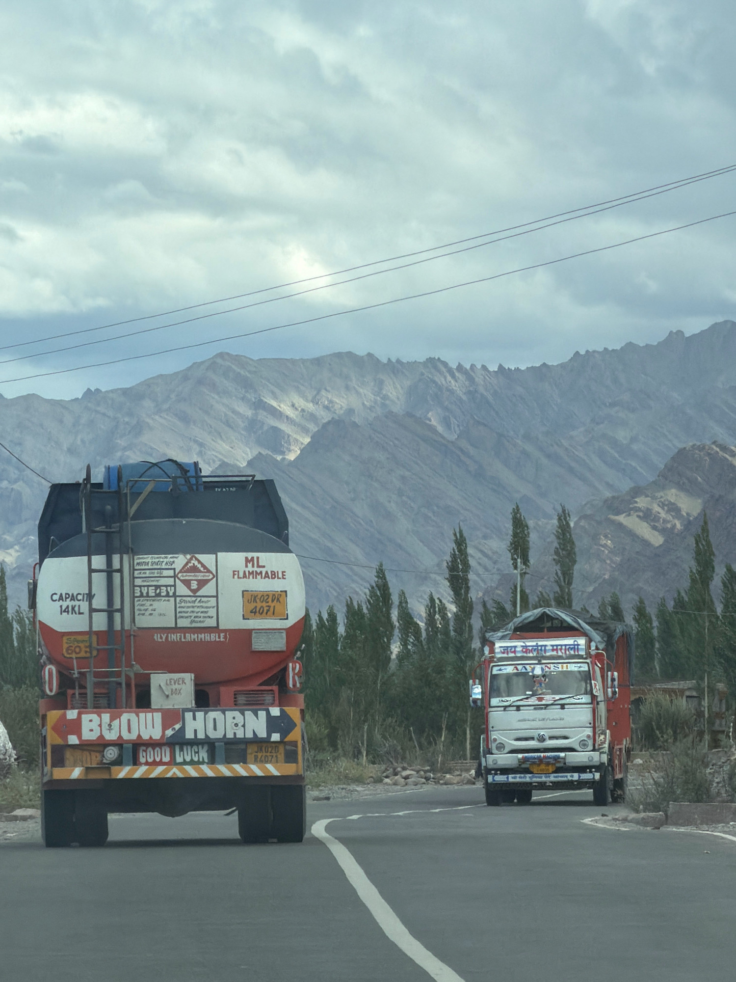 Two trucks are driving on a paved road with a mountainous landscape in the background under a cloudy sky. The truck on the left is a red and white tanker truck with "CAPACITY 14KL" and "BLOW HORN" written on its side and rear. It also has the text "GOOD LUCK" and the license plate "JK02D 84071". The truck on the right is a red and white cab-over truck with Hindi text on its windshield and "Jay Kela Marali" written above it. The environment appears to be a rural area, possibly the city of Yuluk, India, given the presence of similar trucks and license plates. The trees lining the road are slender and tall.