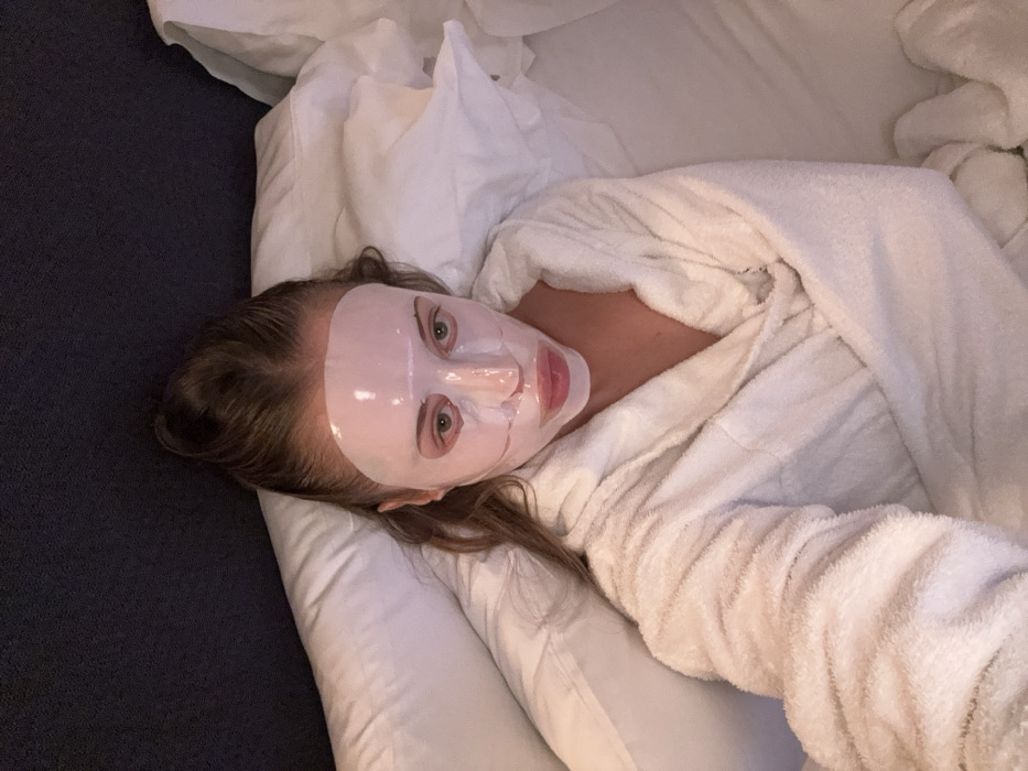 A female subject, approximately 20-30 years of age, is depicted lying on her back in a bed within a residential or hotel bedroom, location Unknown, Unknown. She is wearing a white terrycloth bathrobe and a translucent, light pink facial sheet mask, which covers her entire face from forehead to chin, with cutouts for her eyes, nostrils, and mouth. Her light brown hair is pulled back from her face, with some strands visible around her temples and shoulders. The subject's eyes are open and directed towards the camera. She is positioned on white bedding, which includes pillows and a duvet. A dark grey or black upholstered headboard is visible behind her head. The lighting is low and ambient, consistent with an indoor evening setting. The image appears to be a selfie, captured from an overhead perspective.
