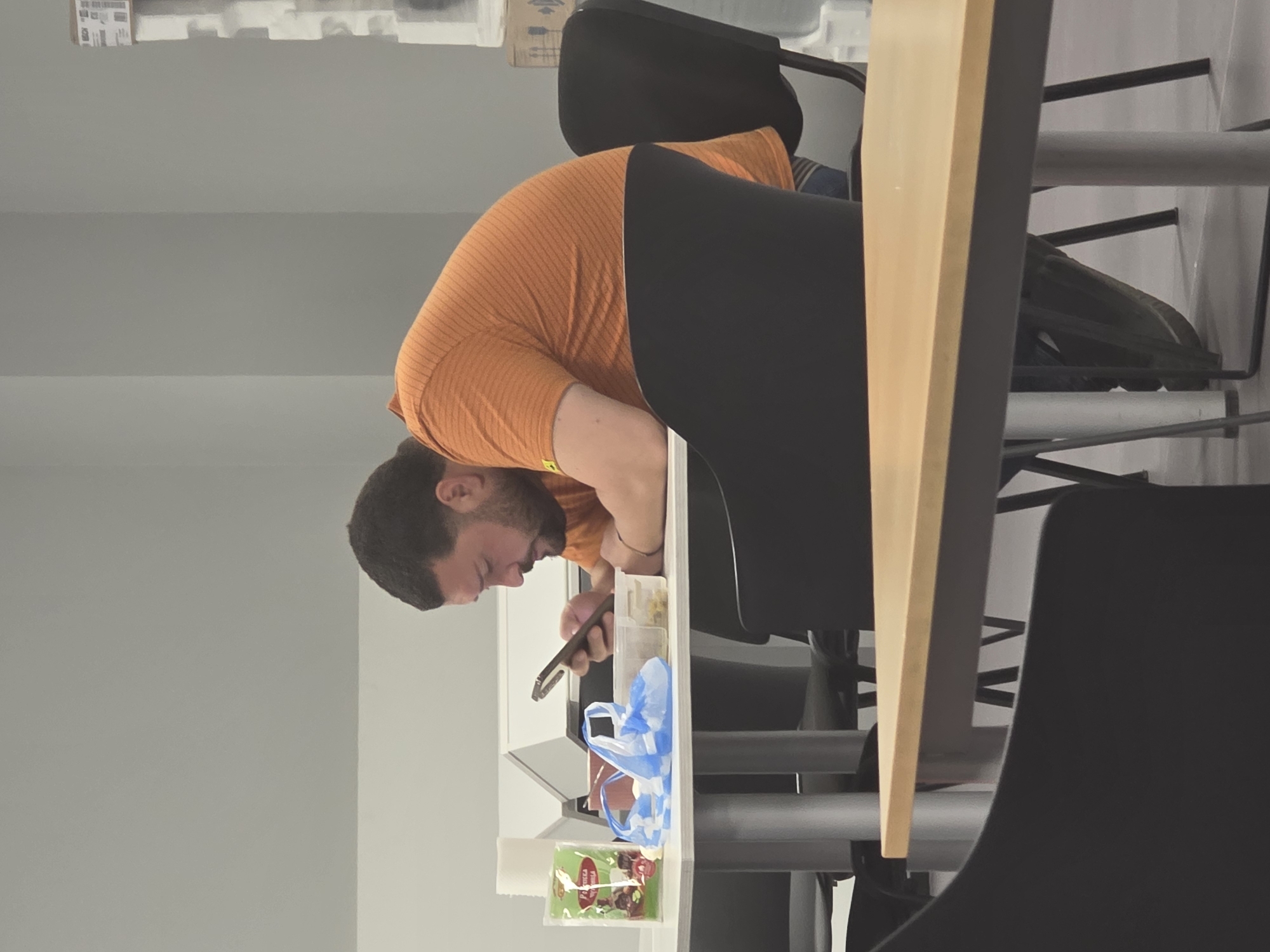 A man with dark hair and a beard is seated at a table, looking down at his mobile phone. He is wearing an orange long-sleeved shirt. On the table are a few food items, including a plastic bag with blue handles, a container of food, and some packaged food. The table is surrounded by black chairs with silver legs. The location is likely an office or cafeteria setting, but there is not enough information to identify the specific location in Plovdiv, Bulgaria. The man's action is using his mobile phone, and the overall environment is indoor and looks like a business cafeteria or office.