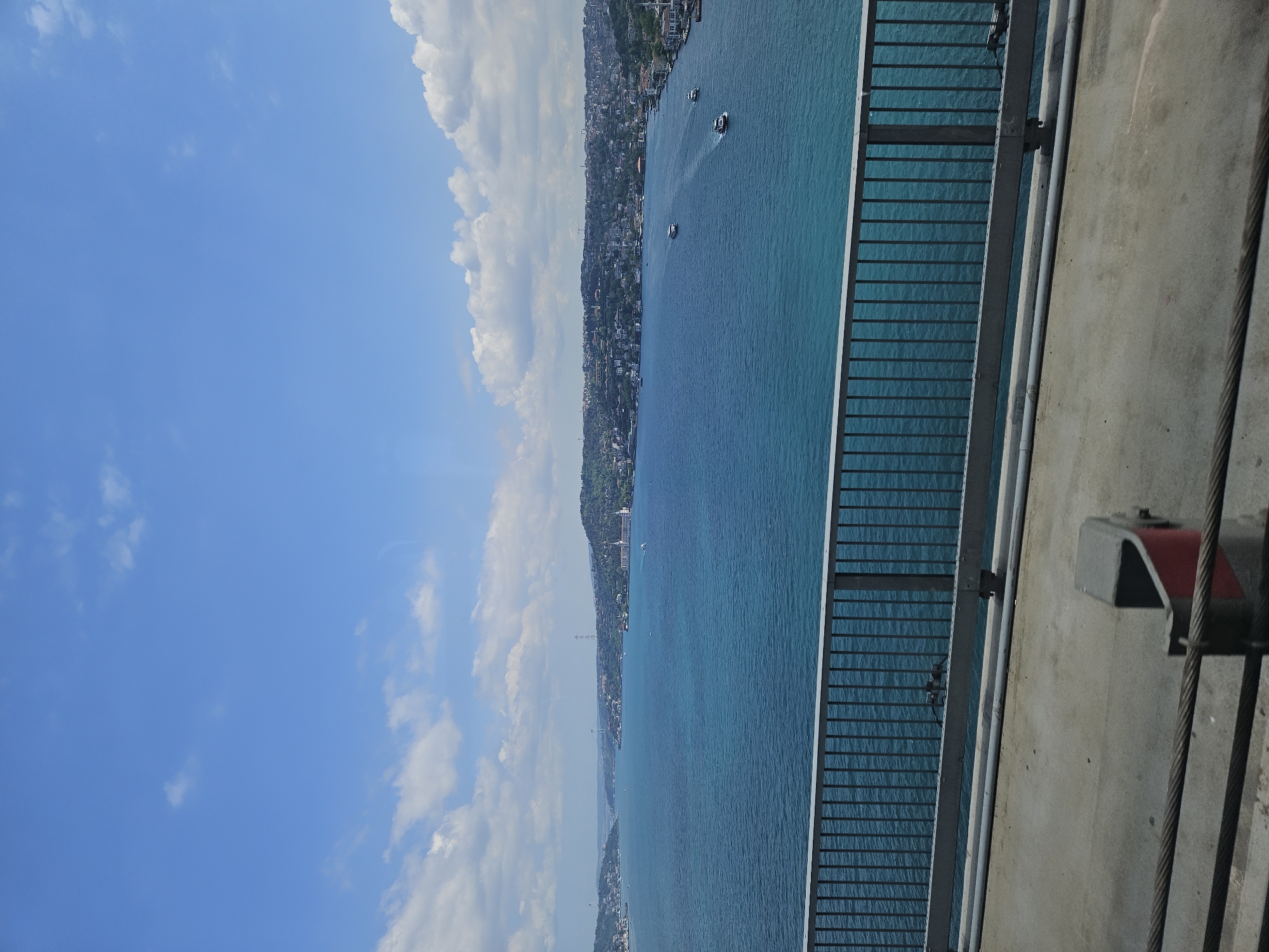 View from the Bosphorus Bridge in Beşiktaş, Turkey. The sky is blue with white clouds. A coastline with buildings and trees runs along the left of the frame, adjacent to a body of blue water with a few small boats. A metal railing and part of the bridge structure are on the right.