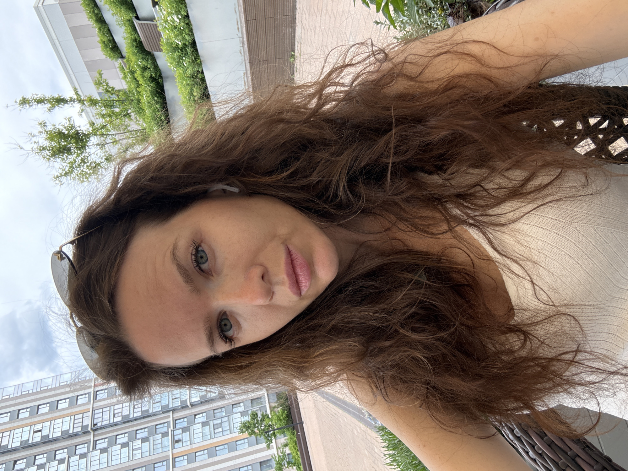 A woman with long, wavy brown hair and light blue-green eyes is featured in a close-up, angled outdoor selfie. Her head is tilted, and she looks directly into the camera with a neutral to slightly soft expression, her lips tinted with light pink lipstick. Sunglasses are perched on top of her head, and a white earbud is visible in her left ear. She has fair skin and is wearing a light cream or beige ribbed knitted top, with part of her arm and shoulder visible.
The background depicts a modern urban environment, consistent with the provided location context of Moscow, Russia. To the left, a contemporary high-rise building with a grid pattern of windows and light-colored panels stands against a bright, partly cloudy sky. Above her head and slightly to the right, another building features a distinctive green facade, appearing to be covered in climbing plants or a vertical garden, with a wooden-textured section. Green trees are scattered throughout the scene, adding natural elements to the cityscape. A paved area is visible to the right, suggesting a walkway or patio, and part of a dark, woven chair can be seen in the lower right corner, indicating she is seated. The scene is brightly lit, suggesting it is daytime. There is no visible text in the image.