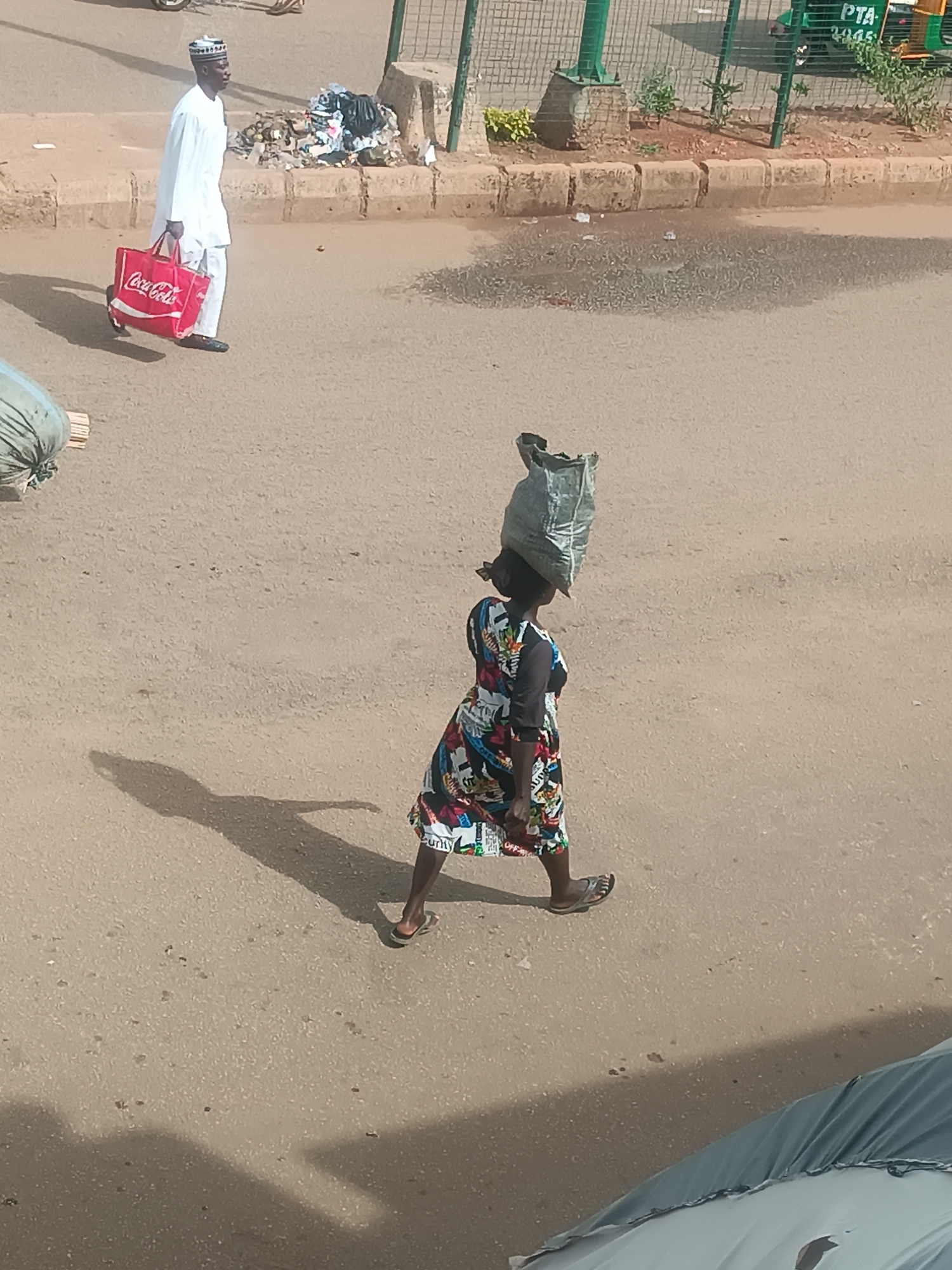 The image captures a street scene in Bukuru, Nigeria. A woman is walking across the street, carrying a large gray sack on her head. She is wearing a colorful dress with black sleeves and flip-flops. Her shadow stretches out on the ground to her left. In the background, a man wearing a white outfit and cap is walking in the opposite direction, carrying a red Coca-Cola bag. Behind him is a pile of trash on the side of the road near a green metal fence and vegetation. A green and yellow auto-rickshaw is partially visible behind the fence. The street surface is a light brown color. The light suggests it is daytime.