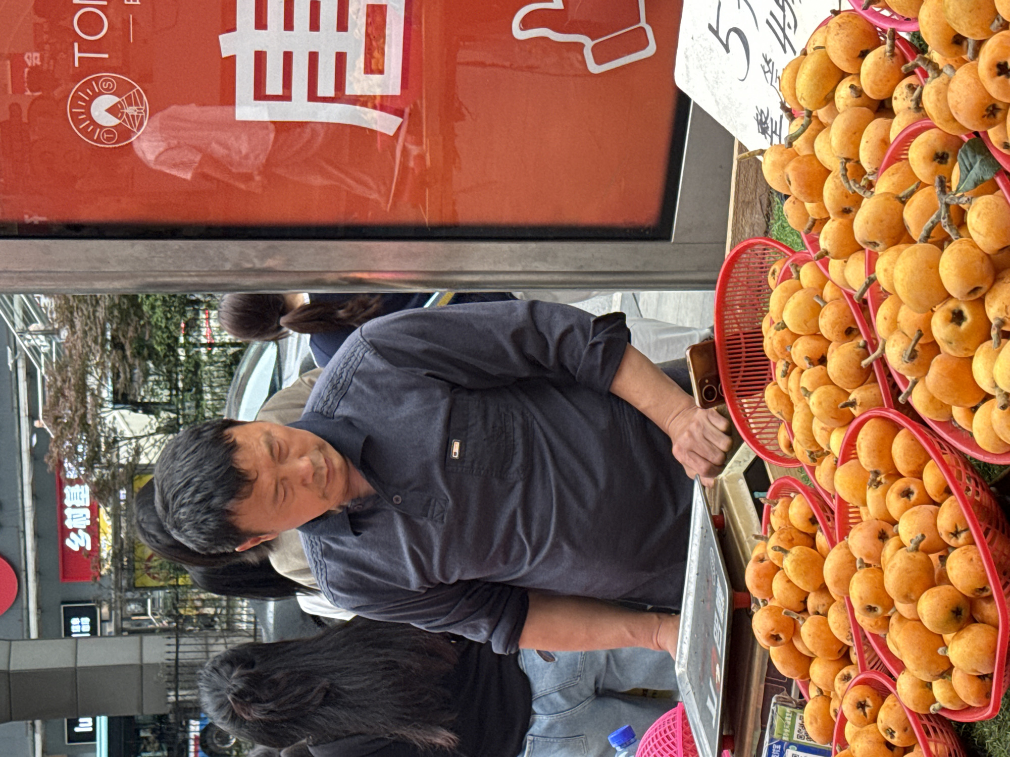 A man in a navy shirt is selling baskets of loquats at a street market in Nan'an District, China. The fruit is displayed in red plastic baskets. The street scene includes other pedestrians, street signs in Chinese, and a food or product advertisement in the background.