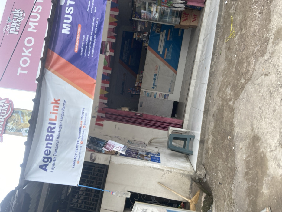 The image shows the storefront of a small shop in Baleendah, Indonesia. The shop is located at coordinates -6.996 latitude and 107.631 longitude. The storefront is adorned with several signs, including one that reads "AgenBRILink Layanan Transaksi Keuangan Tanpa Kantor" and another that reads "Toko Musti." The shop offers various financial services, including cash withdrawals, deposits, and bill payments. The shop is open and there is a counter with a sign that reads "AgenBRILink" above it. There is also a broom and a bucket of water outside the shop. The shop appears to be open for business.