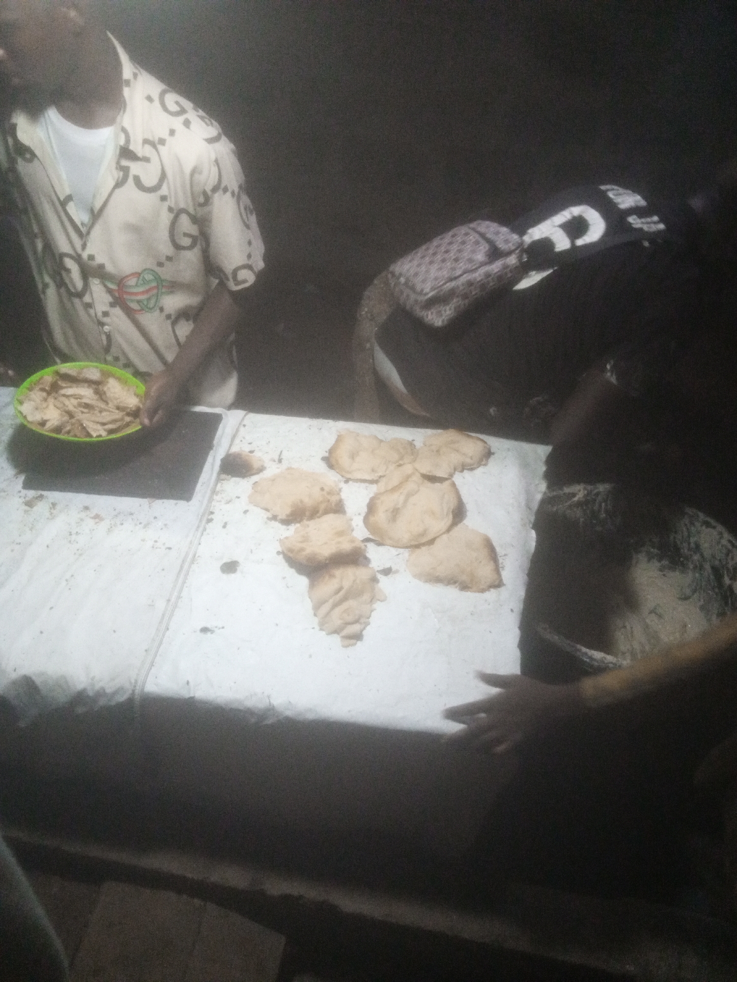 In Tundun Wada West, Nigeria, a dimly lit scene depicts a food stall or gathering. A person wearing a light-colored shirt with a repeating "G" pattern and a red and green embroidered emblem is standing on the left, holding a green bowl filled with light brown, flat, irregularly shaped food items. To the right, a person in a dark shirt with a large white "G" logo is visible, with a patterned bag placed on their lap or chest. A white cloth covers a table or surface where several more pieces of the same light brown, flat food are laid out, some scattered with crumbs. The environment appears to be outdoors at night, with limited light sources, creating shadows and obscuring finer details of the surroundings. The focus is on the food items and the hands of individuals preparing or handling them.