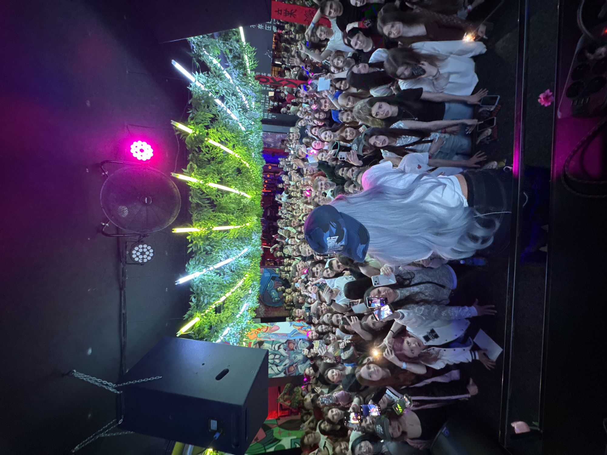 The image depicts a large crowd of people, mostly young, indoors in what appears to be a concert venue in Komsomolsk-on-Amur, Russia. A performer with long white hair, facing the crowd, is likely on stage. Many audience members are holding up their phones, presumably recording or taking pictures. The background features greenery with integrated glowing light bars, possibly a decorative stage element. A black speaker cabinet hangs from chains on the left. A spotlight and a fan are visible on the ceiling. The overall lighting is a mix of pink, green, and yellow, contributing to a concert-like atmosphere. A graffiti-style mural is partially visible in the background.