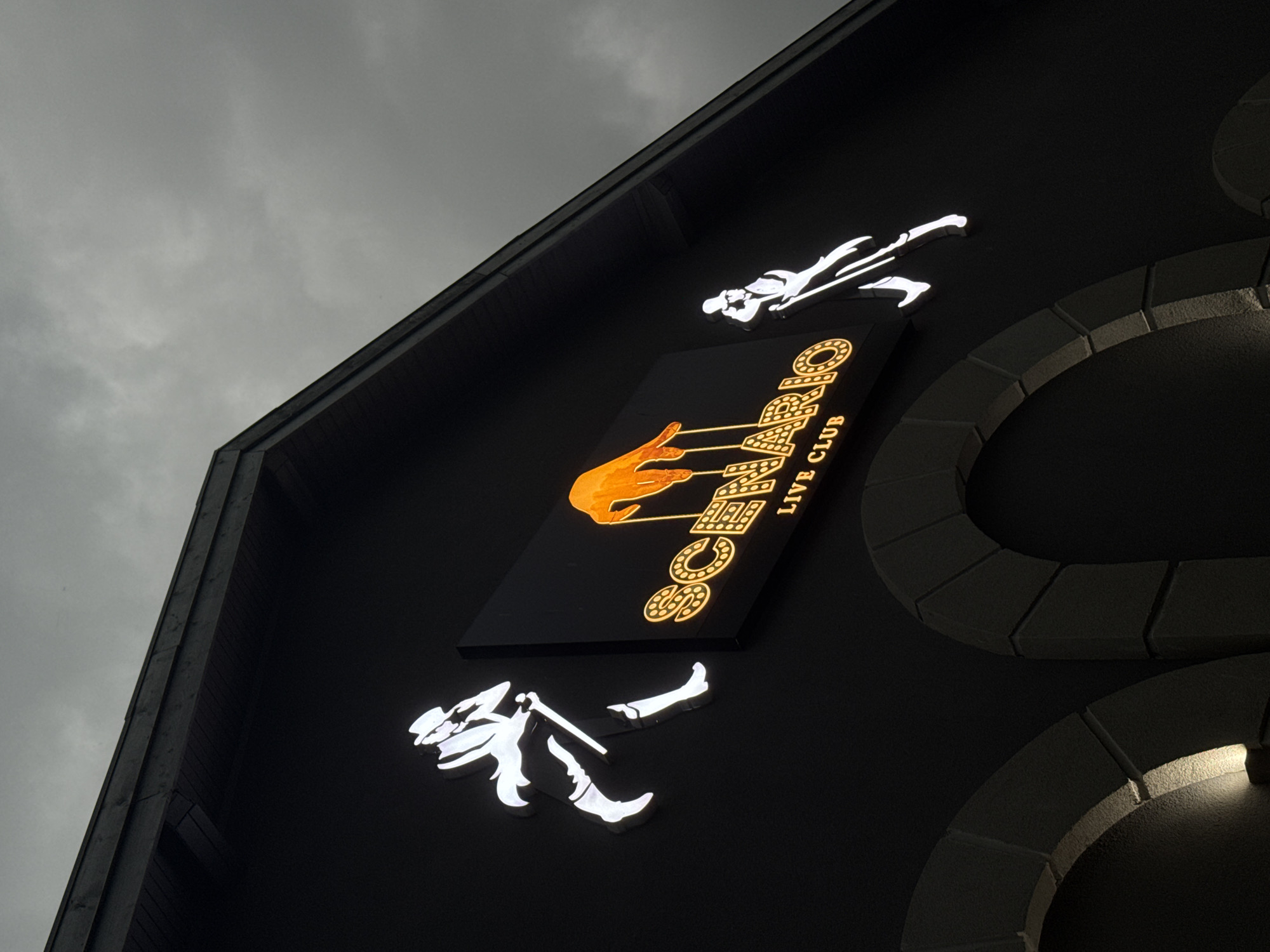 The image presents an upward-angled view of the exterior of a building, identified as "SCENARIO LIVE CLUB" located in Tsarevo, Bulgaria, against a grey, overcast sky. The central subject is a prominent rectangular sign on the dark-toned building facade.
The sign features the club's name, "SCENARIO LIVE CLUB," in bright orange, illuminated text. The word "SCENARIO" is displayed in a larger font, with the letter 'O' designed with internal dots. "LIVE CLUB" is written in a smaller font directly below it. An illuminated orange hand is depicted on the left side of the "SCENARIO" text, with thin lines extending from its fingertips, suggesting the playing of an instrument or strings.
Two stylized, illuminated figures, rendered in white, are positioned above and below the main sign. These figures are depicted in dynamic poses, suggesting dancers or performers. The upper figure appears to be in motion, possibly running or lunging, while the lower figure is in a more crouched or leaping pose.
The building itself has a dark, possibly black or dark grey, facade. To the right of the sign, decorative architectural elements are visible, resembling segmented, stone-like arches, also in a dark tone, subtly illuminated from below to highlight their textured surface. A dark roofline is visible in the upper left portion of the frame. The cloudy, grey sky indicates either a heavily overcast day or dusk, allowing the illuminated signs and architectural lighting to stand out prominently. The scene suggests an entertainment venue or nightclub.