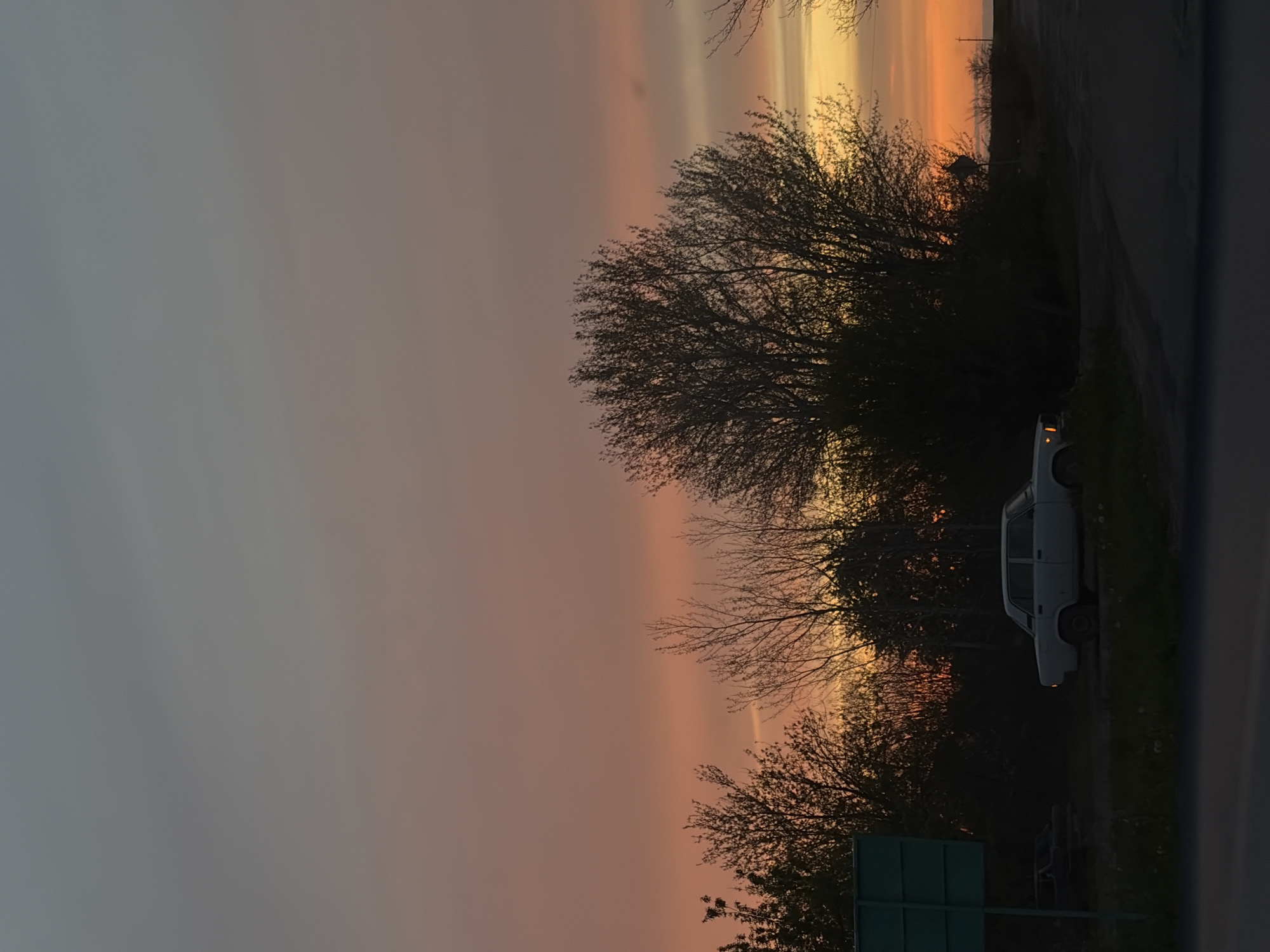 The image, taken in Alexandrovka, Russia, captures a serene moment at either sunset or sunrise. The sky dominates the upper and left parts of the frame, displaying a striking gradient of colors. It transitions from a soft, muted grey-blue on the far left to warmer hues of pink, vibrant orange, and bright yellow on the right, indicating the low angle of the sun.
Dense clusters of trees, primarily consisting of bare, intricate branches, are silhouetted against this colorful sky, occupying the right side of the composition. A dark road or unpaved path runs horizontally along the bottom of the image.
Parked on the right side of this path is a light-colored sedan, possibly white or light grey. Its rear section is visible, and one of its taillights appears to be faintly illuminated. Partially obscured by the foreground trees, a dark green rectangular road sign is visible in the bottom right corner, though no text or symbols can be discerned.
No people or explicit activities are visible; the scene conveys a sense of quietude and natural beauty. The clear sky and vivid atmospheric colors are the main focus, suggesting fair weather. The overall impression is one of a peaceful, contemplative landscape at the transition between day and night.
