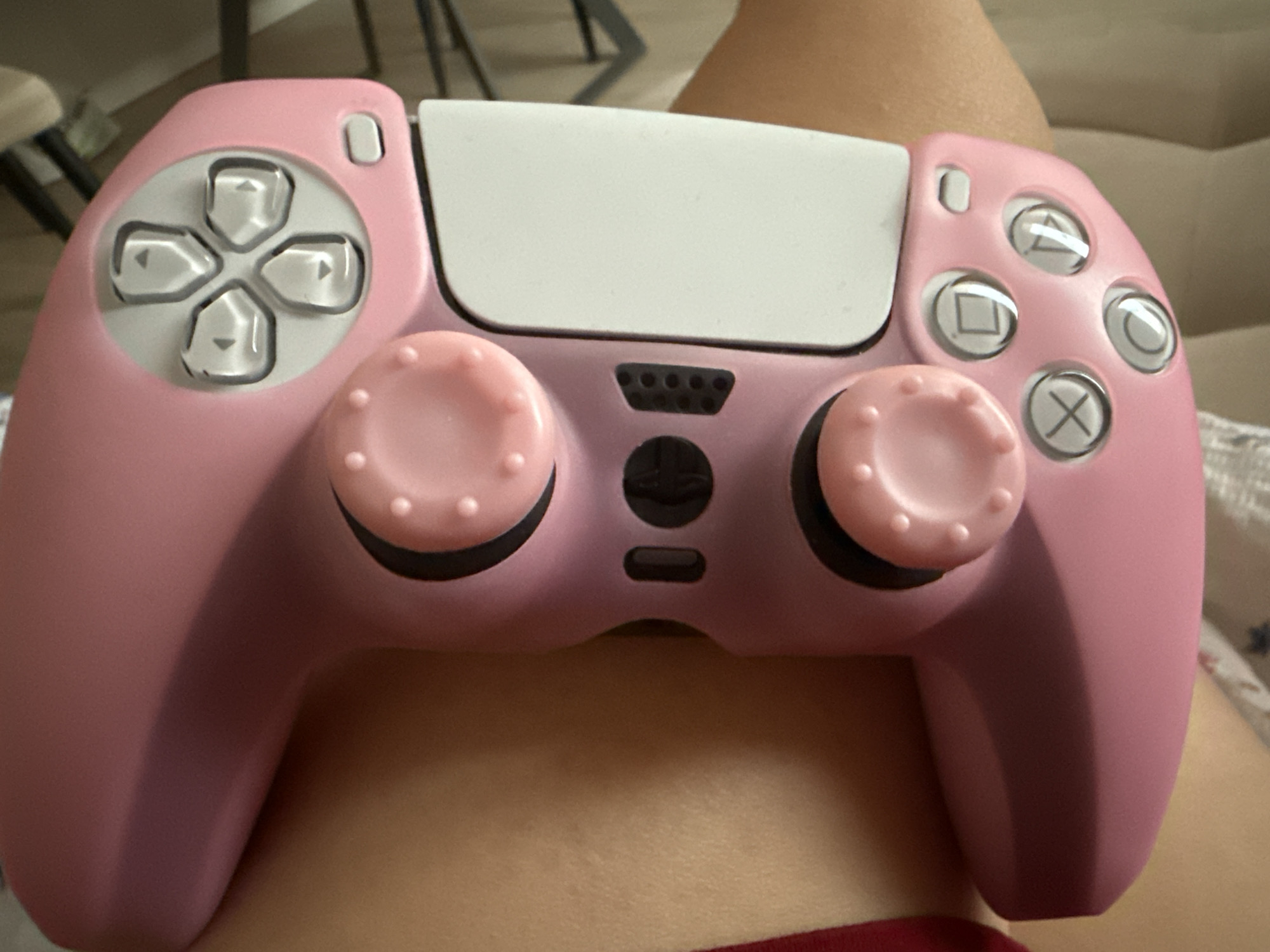A custom pink PlayStation DualSense controller is prominently displayed, resting on what appears to be a person's lap. The controller is a soft, solid pink color with a white central touchpad.
Notable details include the clear, translucent buttons for both the left directional pad (D-pad) and the right action buttons. The D-pad buttons feature silver directional arrows. The action buttons display the standard white PlayStation symbols: triangle, circle, cross (X), and square. Both analog sticks are pink with textured, bumpy tops for enhanced grip.
Other visible features of the controller include small white buttons located near the top left and right corners (likely the 'create' and 'options' buttons, respectively). A speaker grille with multiple small holes is visible below the touchpad, flanked by the two analog sticks. Below the speaker grille, two small, dark buttons are present: a round one in the center (likely the PlayStation button) and a horizontal oval one below it (likely the microphone mute button).
The background is softly blurred, showing a hint of light-toned skin and a piece of red fabric at the bottom, suggesting the controller is on someone's leg or lap. Further in the background, out-of-focus furniture elements can be discerned, indicating an indoor setting. The lighting is soft and even, suggesting an indoor environment, likely during the daytime. No explicit text, apart from the PlayStation symbols, is visible.