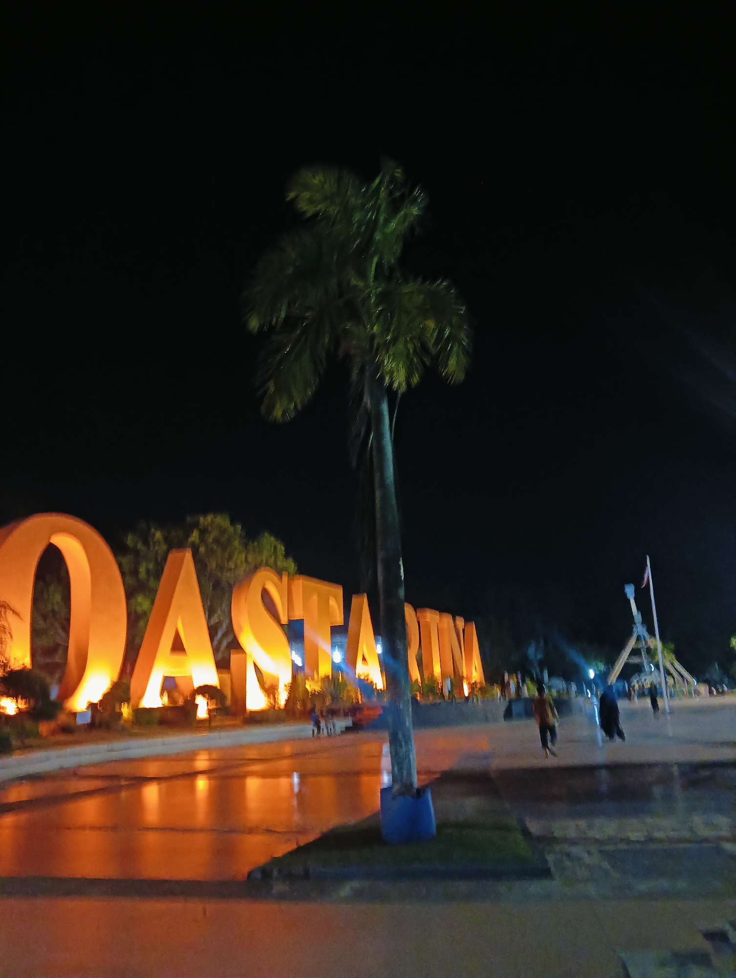 This is a nighttime, outdoor, wide shot of a large, illuminated sign spelling out "DASTAINIMA" in Batam City, Indonesia. The letters are large, orange, and appear to be made of a solid material, possibly concrete or fiberglass. They are lit from below, casting a warm glow on the surrounding area. A tall, slender palm tree stands in front of the sign, obscuring part of the letter "S". The ground in front of the sign is paved and appears to be wet, reflecting the light from the sign. In the background, there are other structures and some people walking. The overall atmosphere is festive and illuminated, suggesting a public space or attraction.