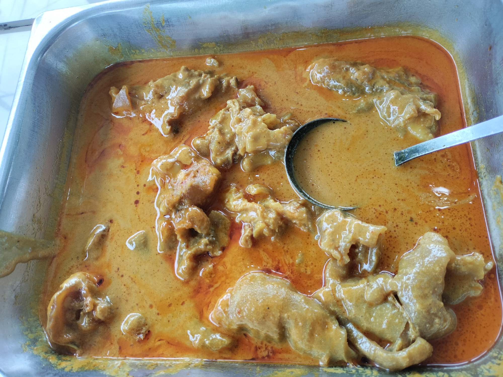 A close-up, overhead shot shows a metal serving tray filled with a rich, orange-colored curry containing large pieces of meat. A ladle with a metal handle is partially submerged in the curry, scooping some of the liquid and meat. The tray is likely in Titibaro, Indonesia, given the context of the request. The lighting suggests an indoor setting, possibly a restaurant or food stall. The curry appears to be a common dish, perhaps chicken or beef, with a creamy sauce. The overall impression is one of a hearty and flavorful meal.