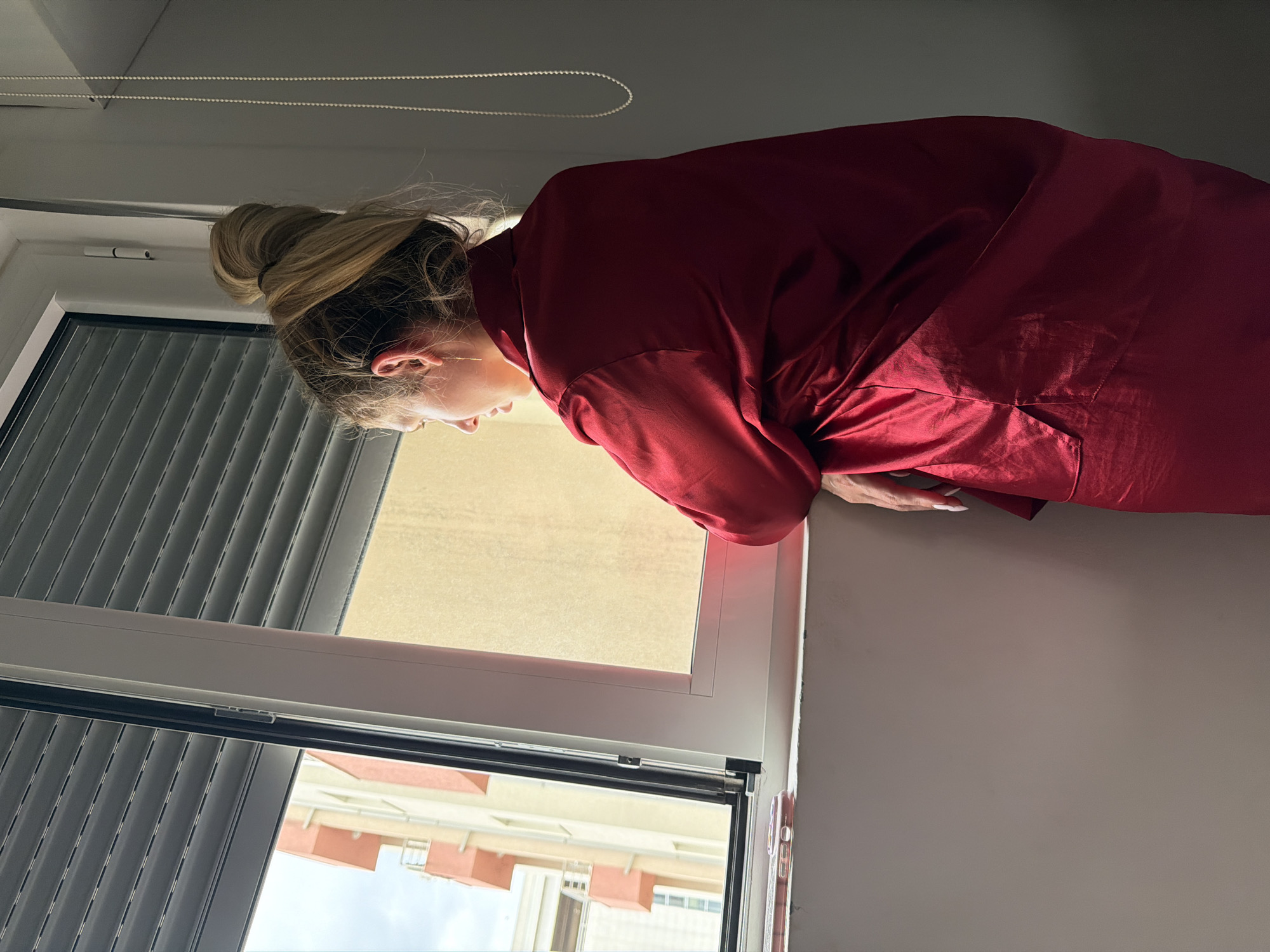 A woman with blonde hair styled in a bun, wearing a red garment, peers out of a window. The window has a partially drawn beige blind and a lower section with horizontal black slats. Outside the window, a multi-story building with light orange facades is visible under a partially cloudy sky. The setting is likely an interior space in an apartment building. While the location is allegedly Budva, Montenegro, there is no explicit visual confirmation within the image itself.