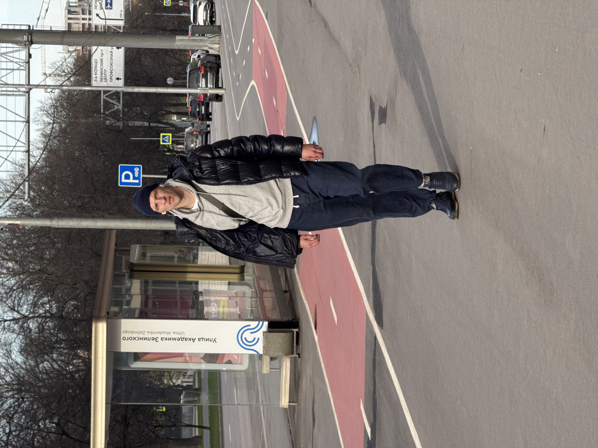 The image shows a person wearing a dark blue beanie, black puffer jacket, gray hooded sweatshirt, dark blue pants, and dark sneakers standing on a paved area, likely a sidewalk or a pedestrian zone, marked with a red lane. The person is facing to the left with a relaxed posture.
The location appears to be a street in Russia. This is indicated by the street signs, one of which says "Ulitsa Akademika Zelinskogo" in Russian and English, along with another sign that includes the words "Leninsky Prospekt" and "Center." A parking sign is also visible, as well as street signs pointing to the "3rd Ring Road." Cars are parked along the road. Buildings and trees are visible in the background. The weather seems overcast.