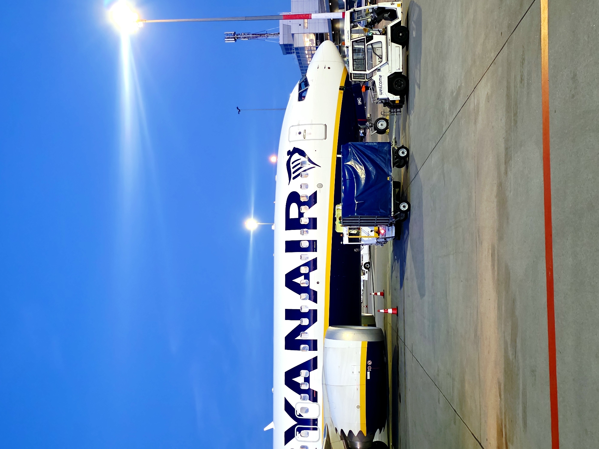 A Ryanair airplane is parked on the tarmac at Poznan Lawica Airport. The plane's fuselage is white with the dark blue "RYANAIR" logo and a stylized wing emblem. A luggage cart covered with a blue tarp is positioned in front of the plane, along with a small white service vehicle that has the word "welcome" printed on its side. Two orange and white traffic cones are placed near the airplane's wheels. In the background, airport buildings and a telecommunications tower are visible. The sky is a deep blue, and a bright light from a lamp post is casting a glare across the scene. The image is taken during twilight.