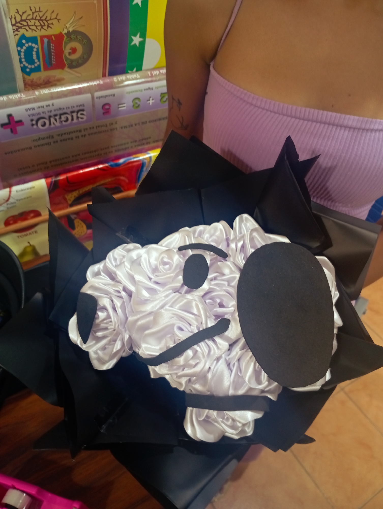 A person wearing a pink ribbed tank top is holding a bouquet of light purple satin fabric roses, arranged to resemble a character. The bouquet is wrapped in black paper. In the background, there are educational posters with colorful graphics, including what appears to be a diagram of a tomato and a pear, and a poster with mathematical equations. The scene is lit by artificial light. The image does not provide enough information to determine the specific location within Juan Griego, Venezuela.
