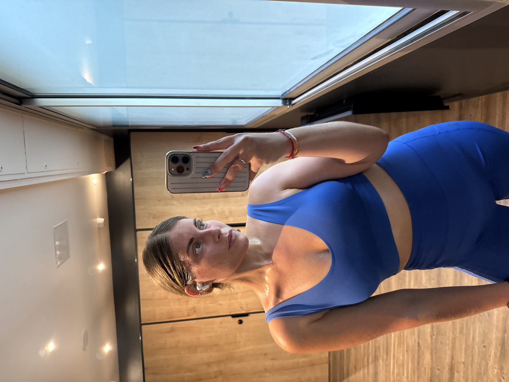 A person is taking a mirror selfie in a gym locker room. The person is wearing a blue sports bra and matching leggings. They have sweat on their skin and are looking to the side. In their right hand, they are holding a phone with a silver case. The background shows wooden lockers and a large window. The image does not contain any information to indicate it is located in Paris, France.