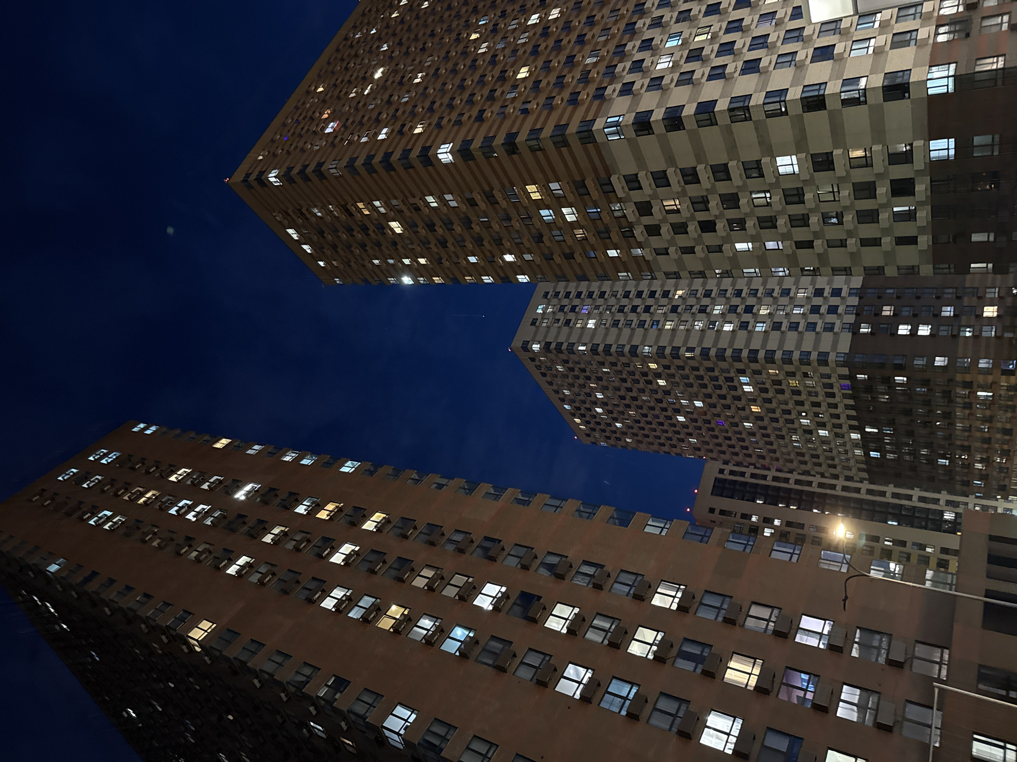 The image captures an upward-looking view of three modern high-rise buildings against a dark blue night sky in Khimki, Russia. The perspective emphasizes the towering scale of the structures, which appear to be residential or office buildings constructed from light-colored materials. Their facades are characterized by a repetitive grid of numerous rectangular windows.
Many of these windows are illuminated, emitting various light colors, including bright white, warm yellow, and cool blue, indicating activity within. Some windows also feature visible external air conditioning units or similar fixtures. A street light with a distinct yellow glow is partially visible in the lower right, contributing to the foreground lighting.
The sky is clear and dark blue, suggesting a cloudless night. A faint, small bright dot, possibly the moon or a prominent star, is discernible in the upper left portion of the sky. The scene conveys a sense of urban nightlife and the quiet majesty of city architecture. No people are visible, and no discernible text is present in the image. The overall impression is one of nocturnal calm in a bustling urban setting.