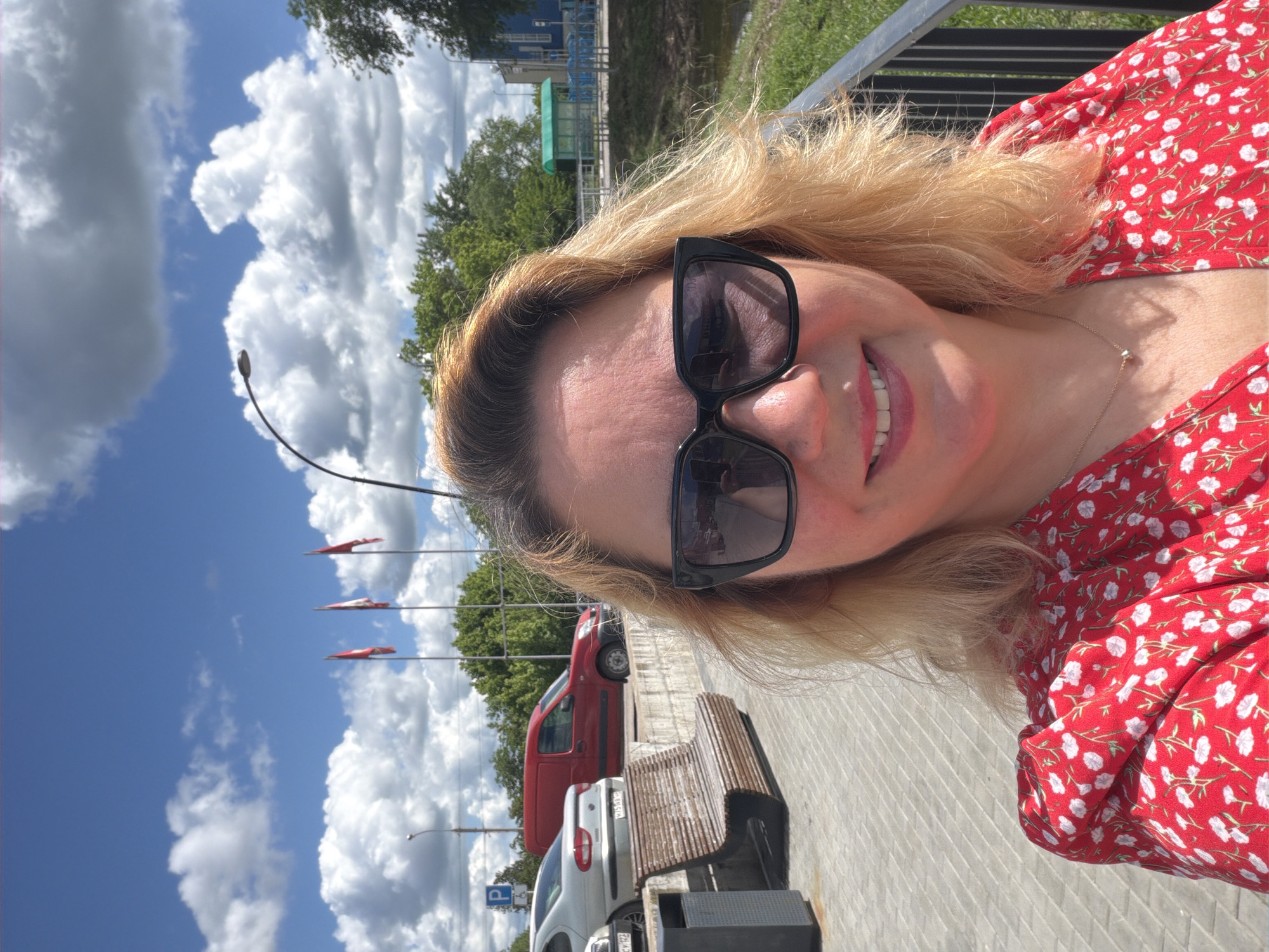 A selfie of a fair-skinned woman wearing sunglasses and a red floral dress is the central focus. The background indicates an outdoor scene in the city of Leningradskaya, Russia. To the left of the woman, three red flags stand tall on poles. Behind the woman, a red delivery van and a white car are parked on a concrete surface beside a weathered wooden bench with a gray metal base. Beyond the vehicles, trees and a blue building are visible. A partially clouded sky is present. The lighting suggests a sunny day.