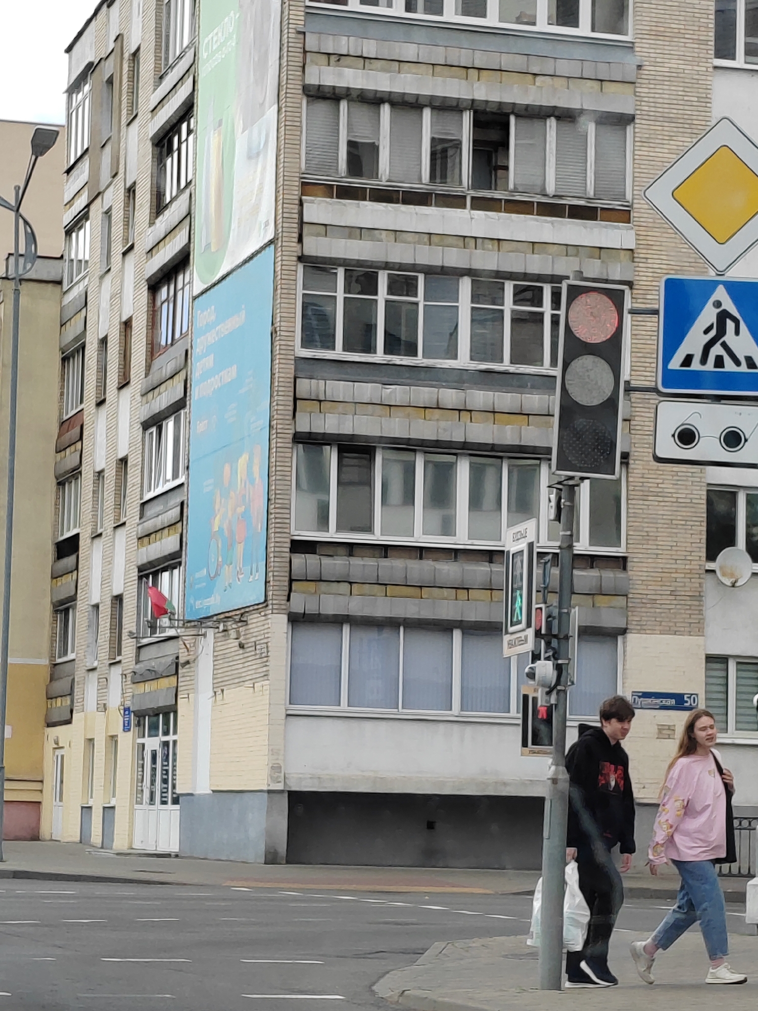 The image depicts a street scene in Brest, Belarus. A multistory apartment building with a light brick facade and grey horizontal banding dominates the background. A large blue banner is affixed to the building's facade, featuring text and images of children. A red flag is visible on a pole extending from the building. A traffic light with a red signal illuminated is present in the foreground, along with traffic signs indicating a pedestrian crossing and a yield sign. Two young individuals, a male in a black hoodie and black pants and a female in a pink sweatshirt and blue jeans, are walking on the sidewalk. The ground is paved, and road markings are visible. The overall environment suggests an urban setting with typical street infrastructure.