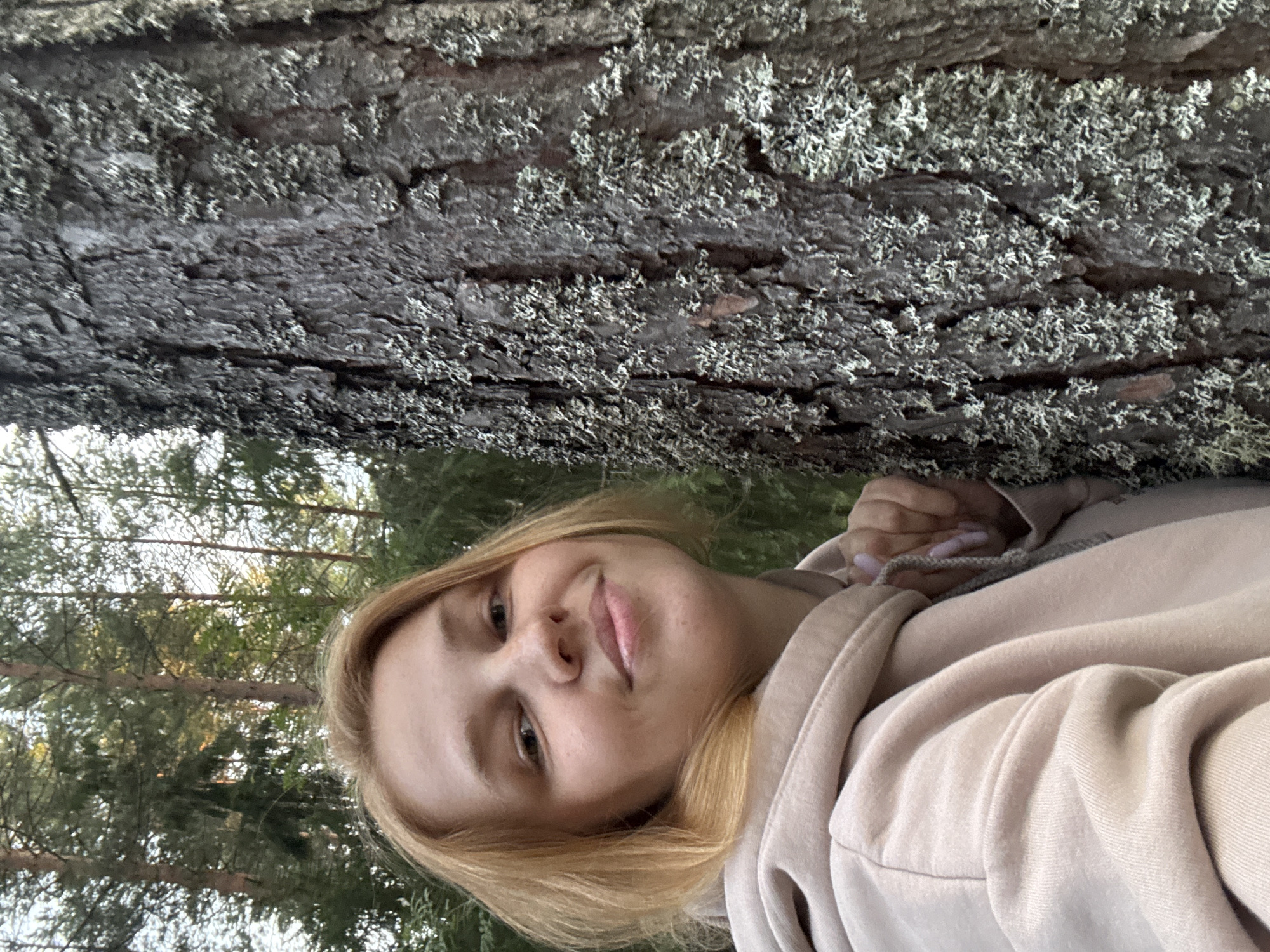 A young Caucasian woman with blonde hair and a light pink hoodie is posing for a selfie next to a tree. She is looking at the camera with a slight smile. The tree bark is rough and covered in light-colored lichen. The background shows a forest with tall trees and green foliage. The scene is set in Anteplevo (Okhotinskoe S/P), Russia.
